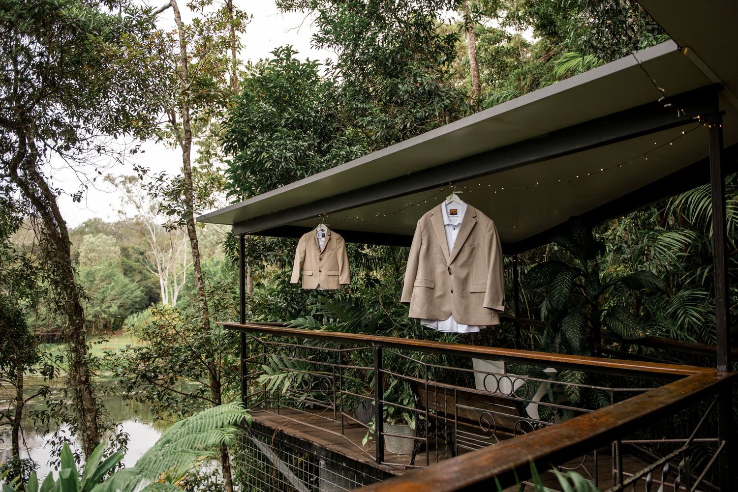Two linen wedding suits hanging on a balcony railing surrounded by lush tropical rainforest greenery.