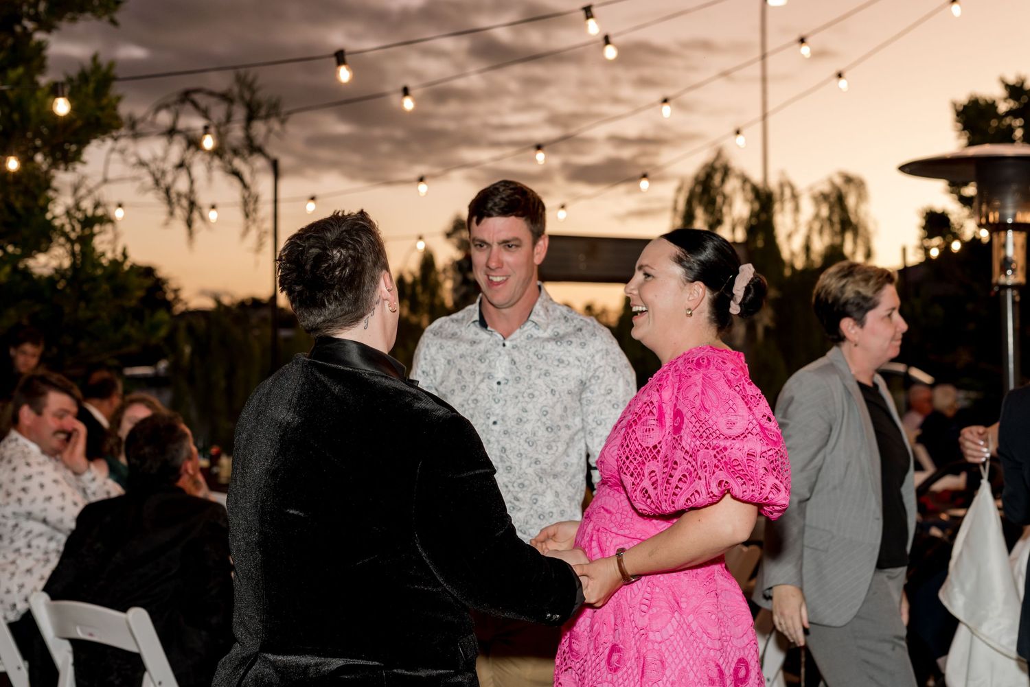 Guests socializing at an outdoor wedding reception under string lights at sunset.