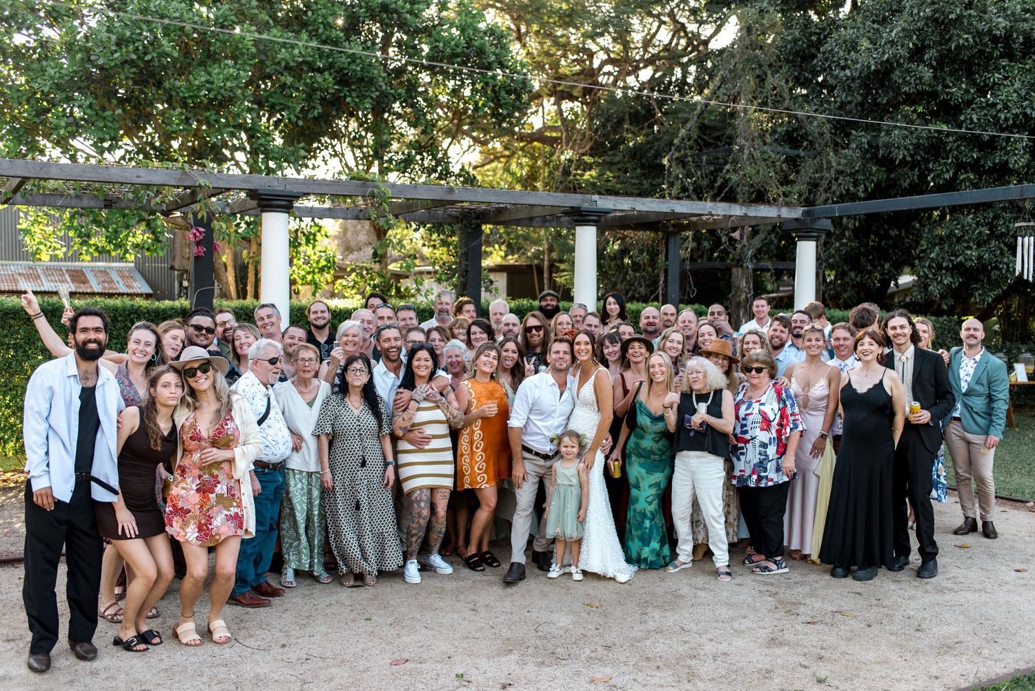 Large wedding group photo with bride and groom surrounded by all guests at outdoor garden venue.