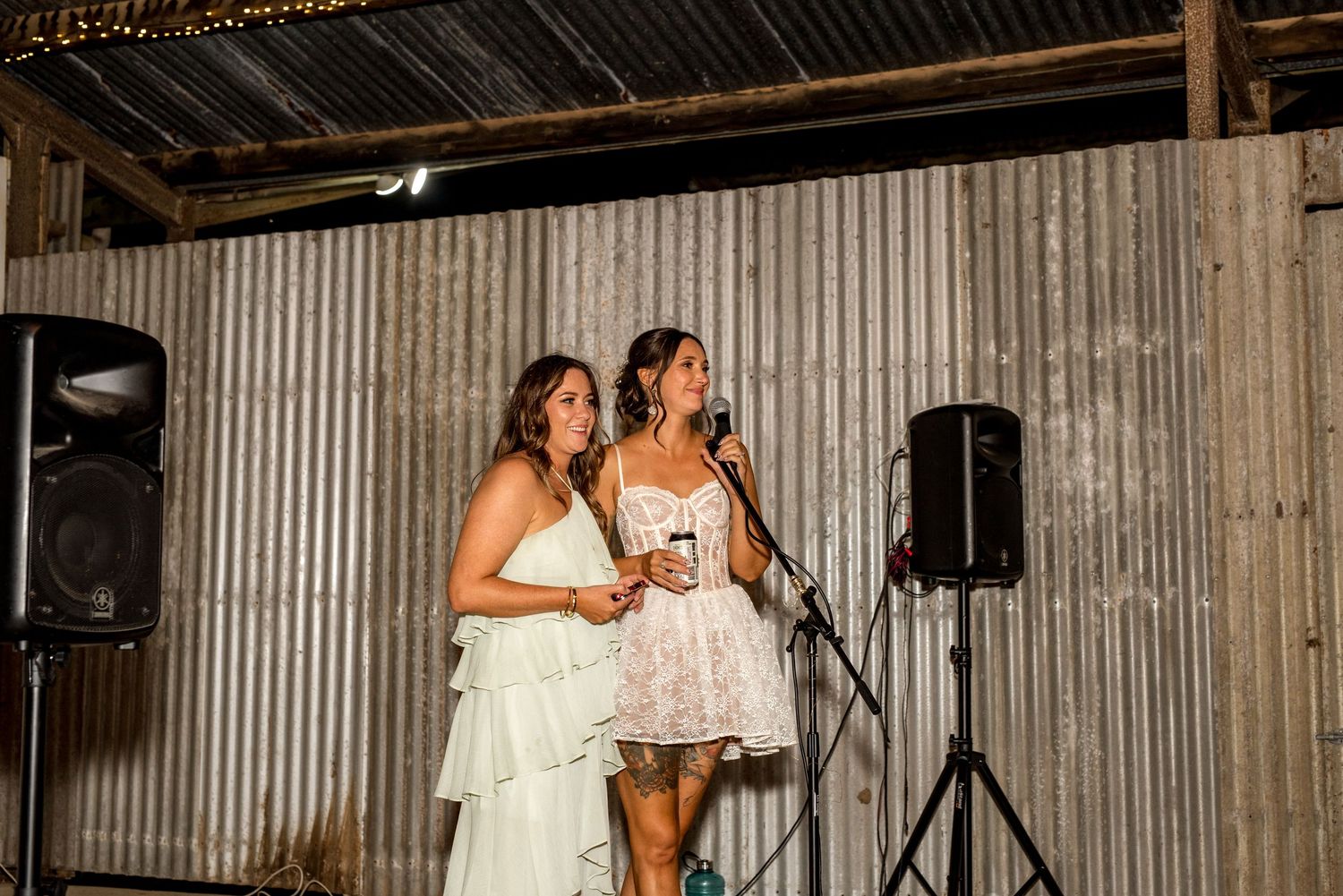 Two women sharing a toast speech at a microphone during a rustic barn wedding reception.