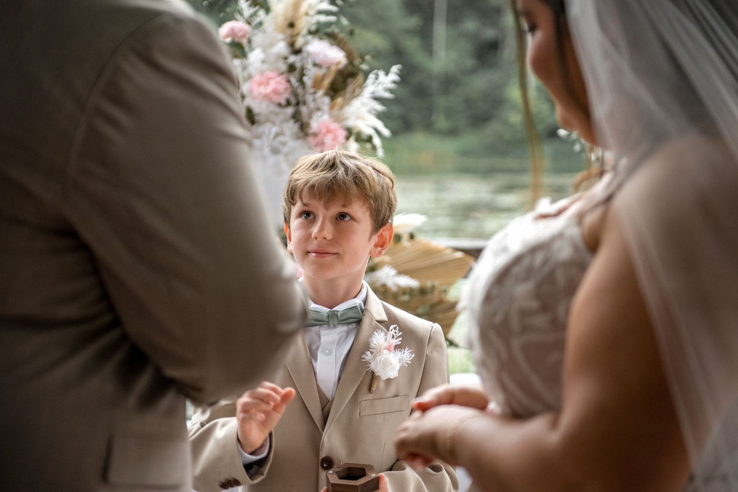 Adorable young ring bearer in matching tan suit looks up at bride and groom during wedding ceremony.