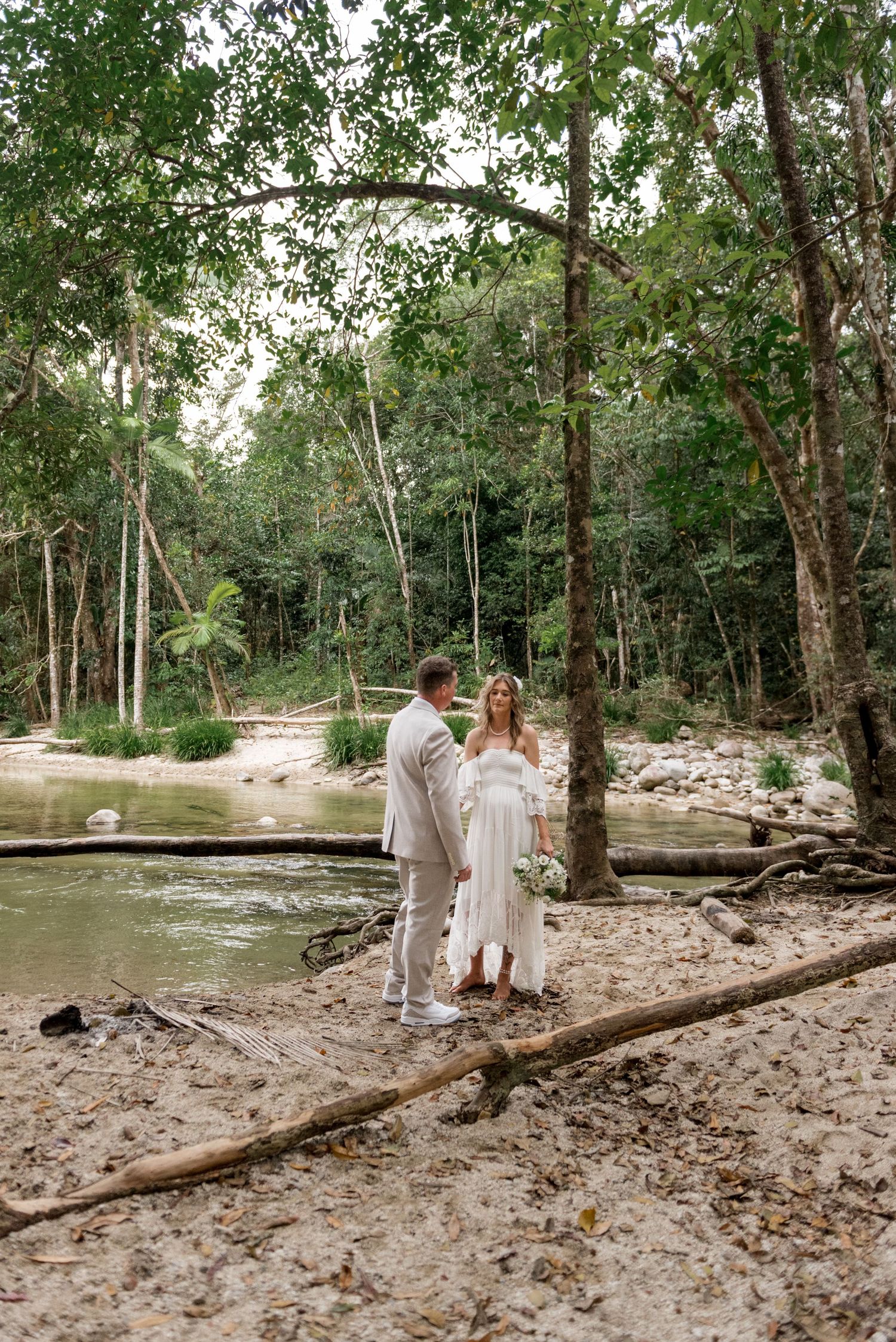 Couple exchanges vows beside a tranquil rainforest river surrounded by tall tropical trees.