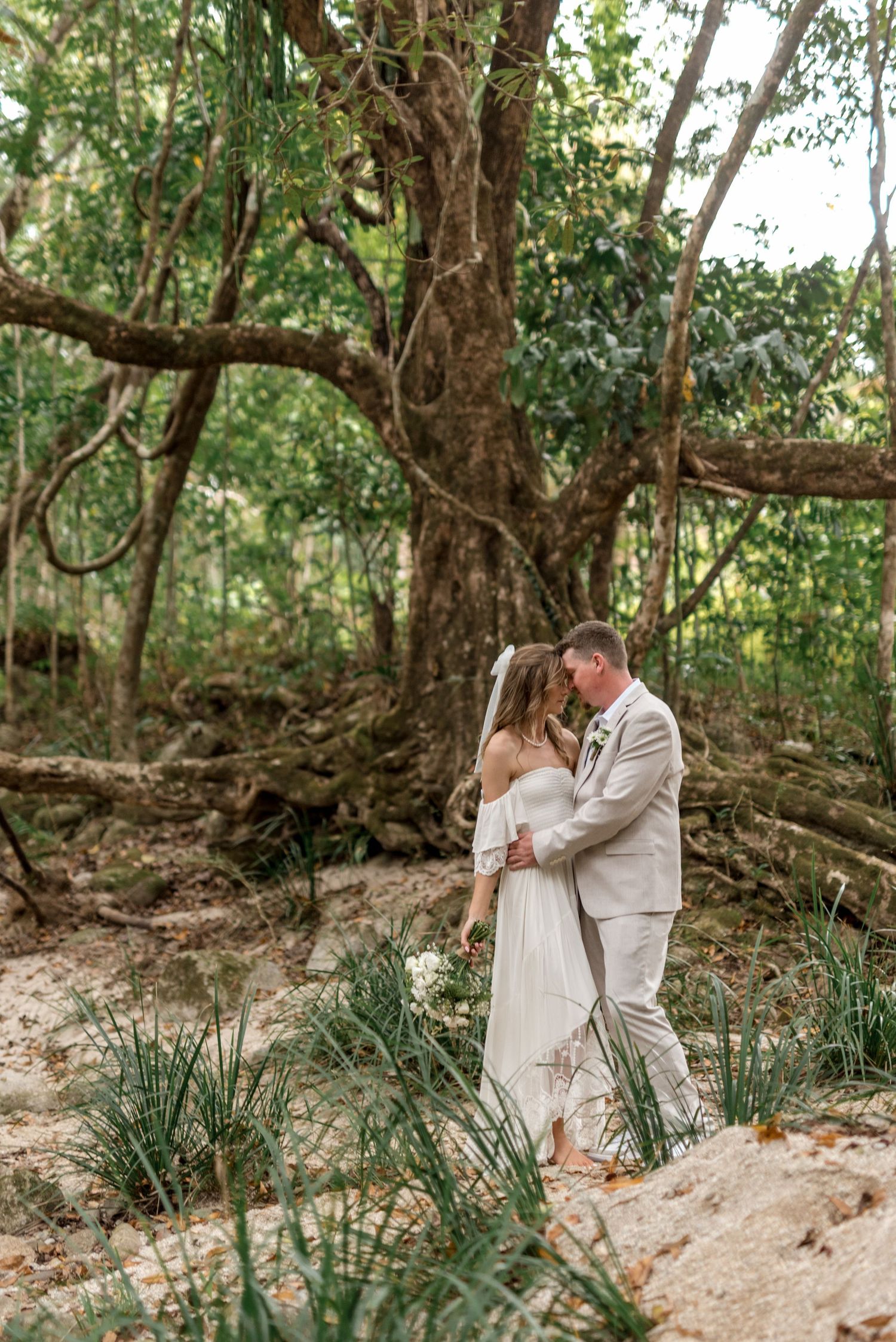 Newlyweds embracing near large tree in tropical forest, bride in off-shoulder white dress, groom in beige suit.