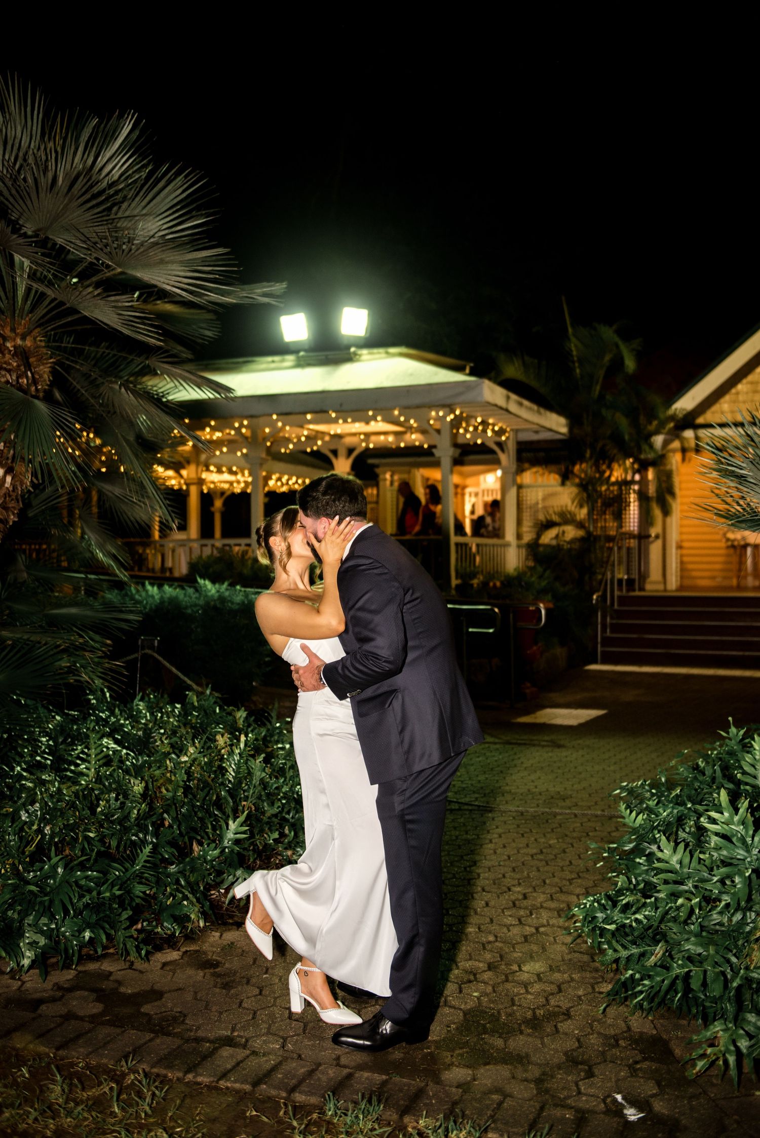 Bride and groom sharing a romantic kiss outdoors at night with fairy lights glowing on a venue behind them.