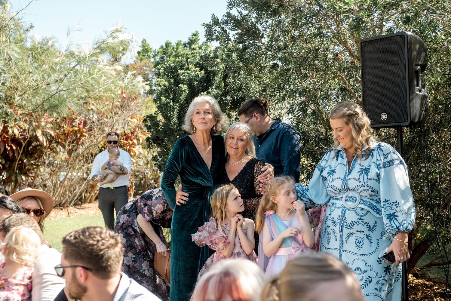 Elegant wedding guests pose together outdoors, including women in teal and floral dresses with young girls in a garden setting.