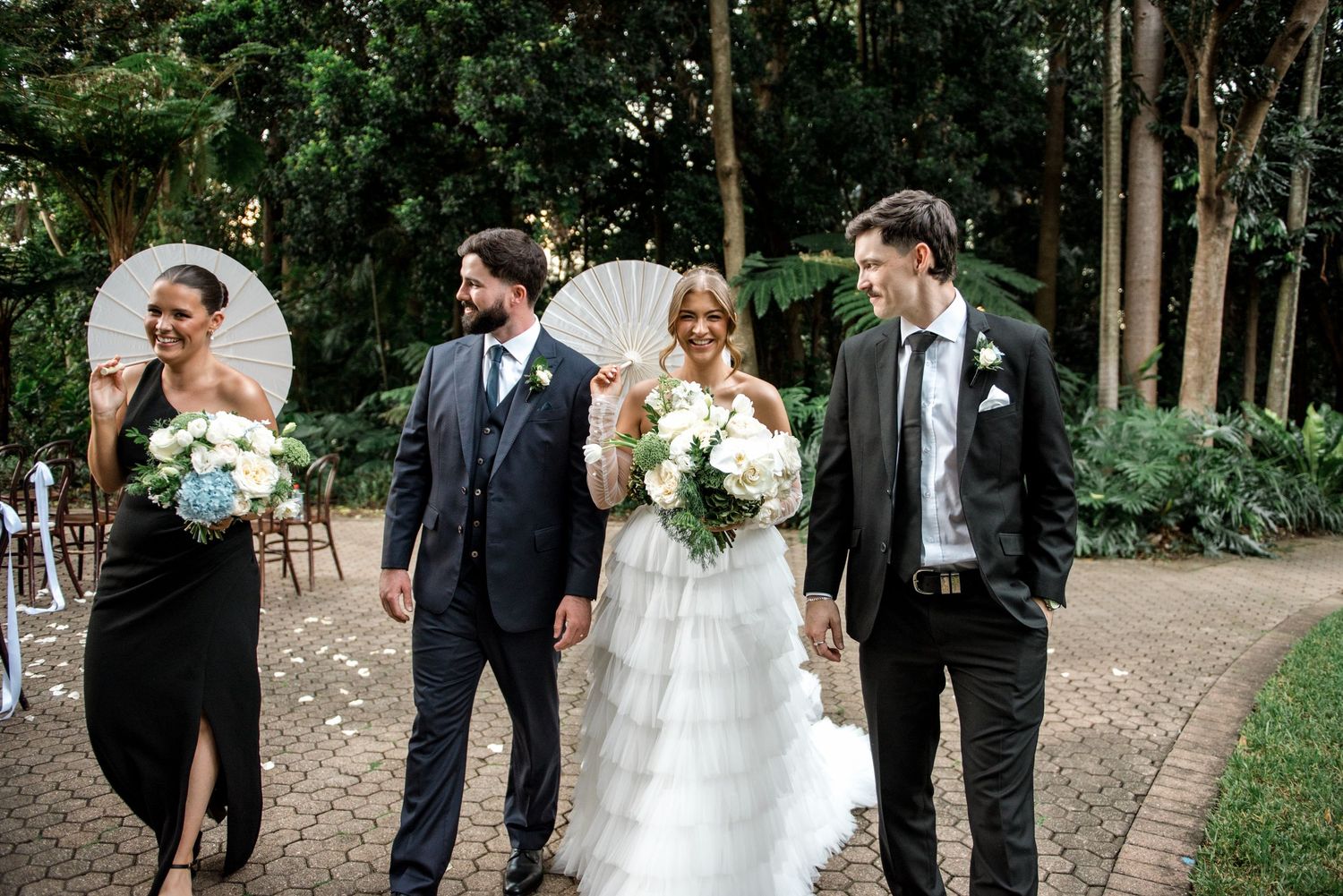 Bride in ruffled gown and groom walk with wedding party holding parasols through a garden ceremony.