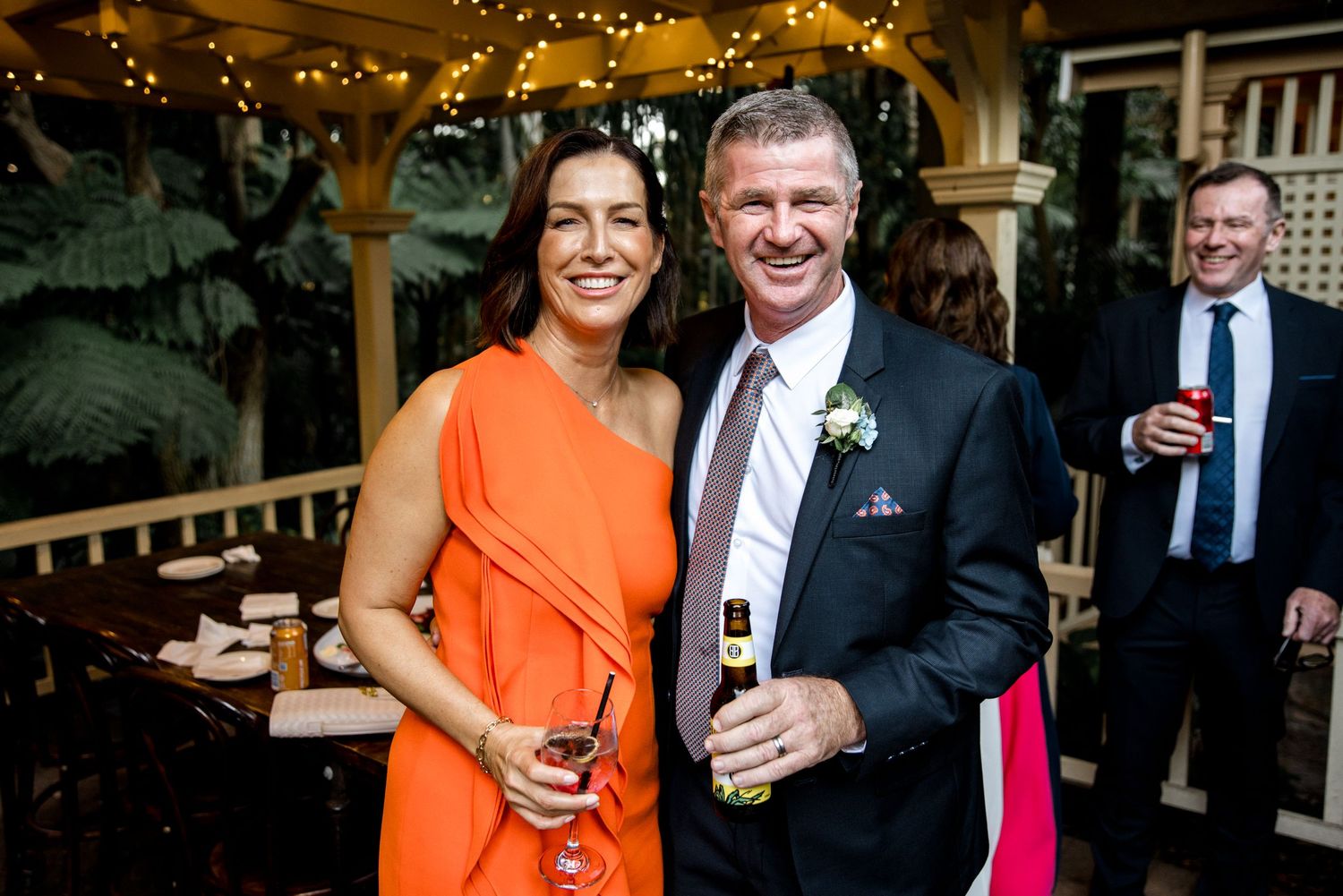 Couple in formal attire smile together at an outdoor wedding reception with string lights.