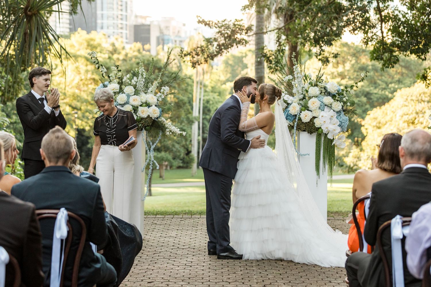 Bride and groom share first kiss at outdoor wedding ceremony with floral arch and seated guests.