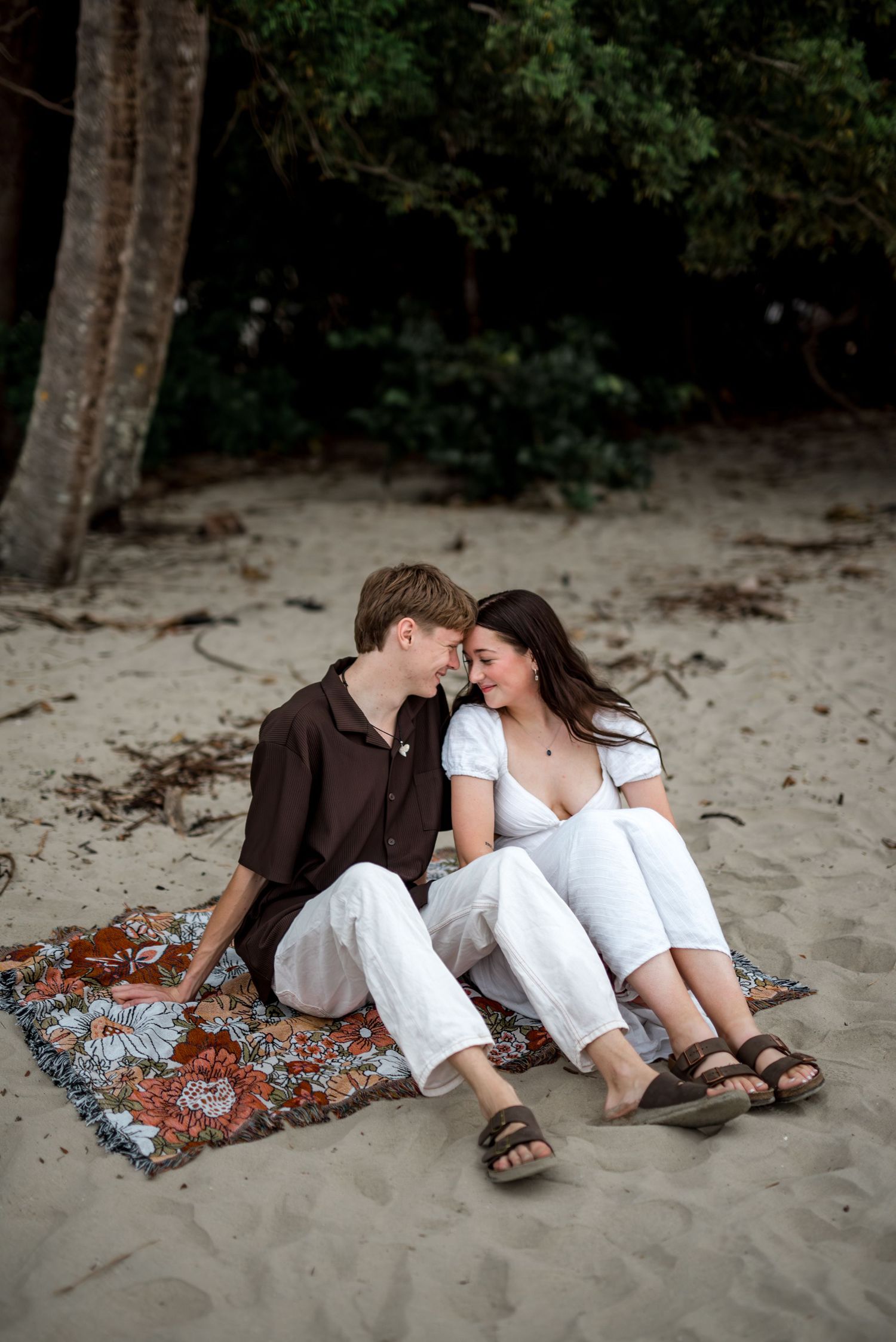 Couple sitting close together on a floral blanket on a sandy beach surrounded by lush green trees.