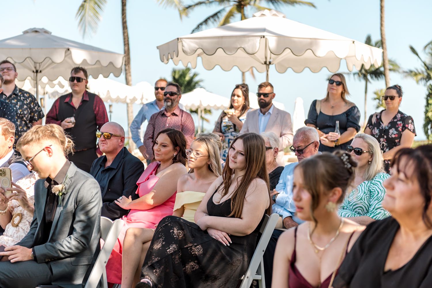 Guests seated in white chairs watch outdoor tropical wedding ceremony under white umbrellas on a sunny day.