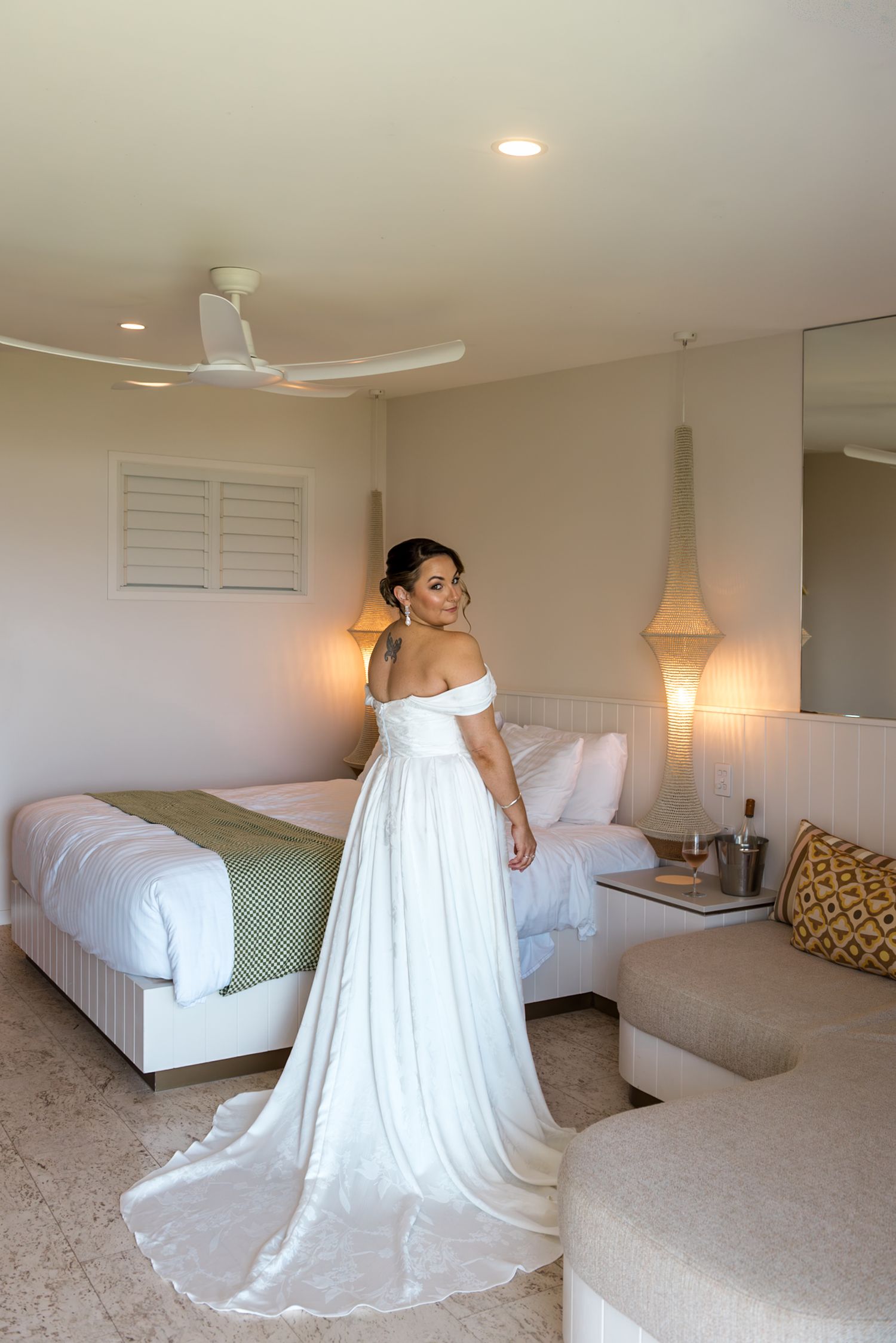 Bride in flowing off-shoulder white gown with long train posing in a bright hotel room.