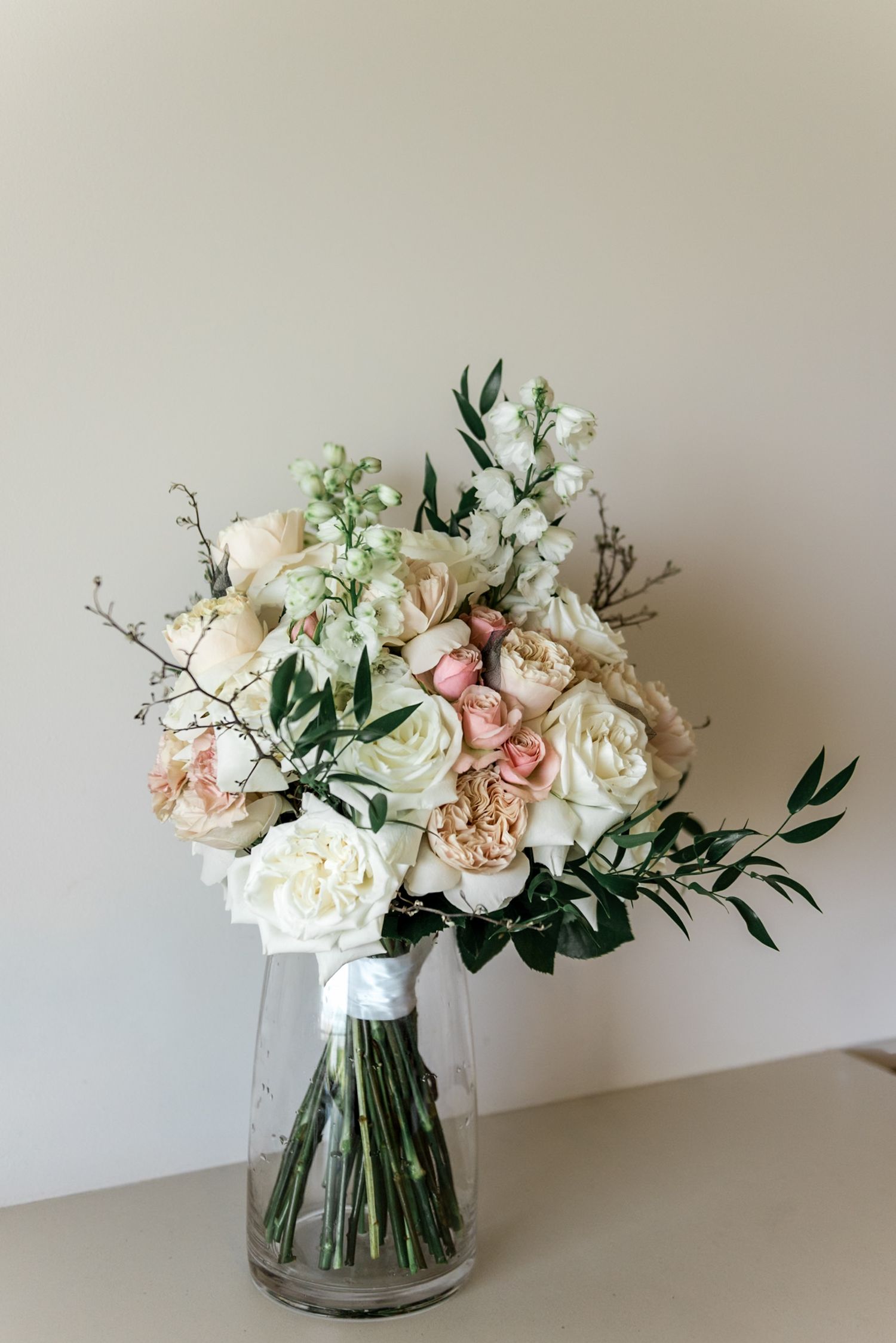Romantic bridal bouquet of white and blush roses with greenery displayed in a clear glass vase.