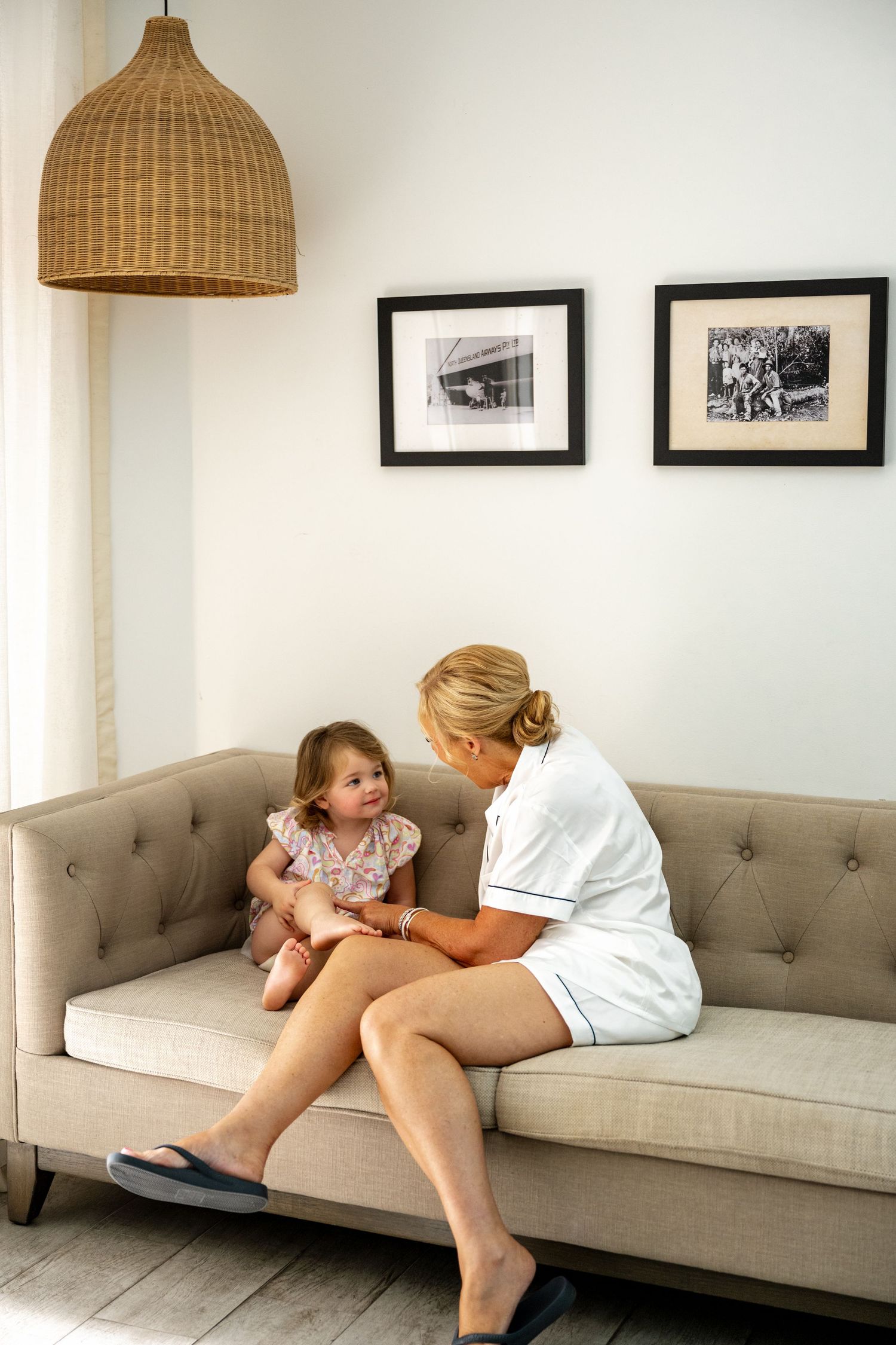 Blonde woman in white dress sitting on a beige sofa interacting with a smiling toddler girl.