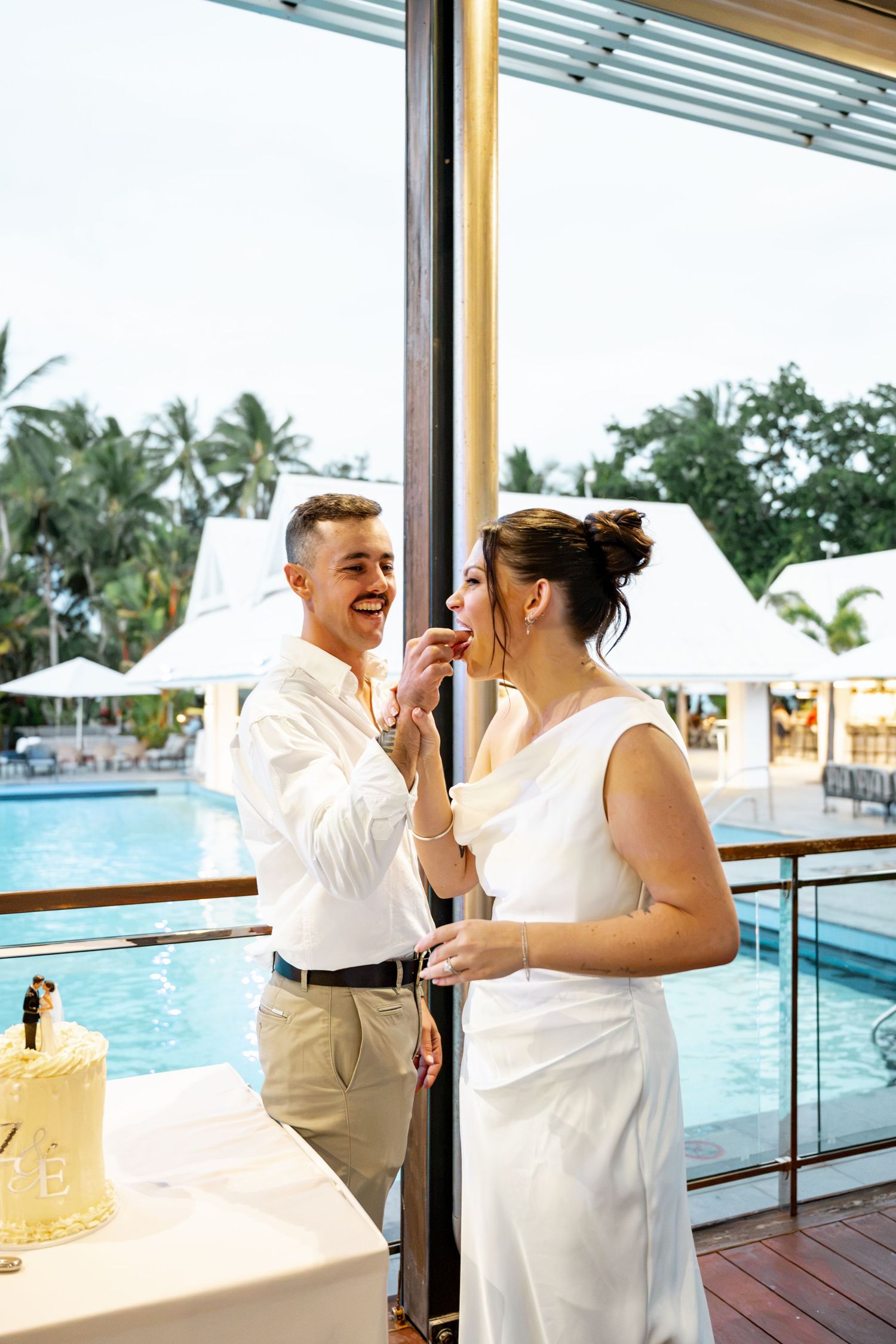 Bride and groom sharing wedding cake beside a tropical resort pool during their reception, Sheraton Grand Mirage Port Douglas.