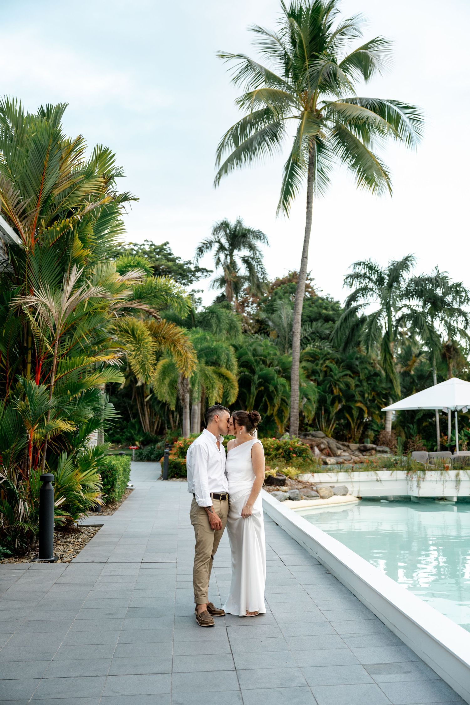 Bride and groom sharing a tender kiss beside a tropical resort pool surrounded by palm trees at Sheraton Grand Mirage Port Douglas.
