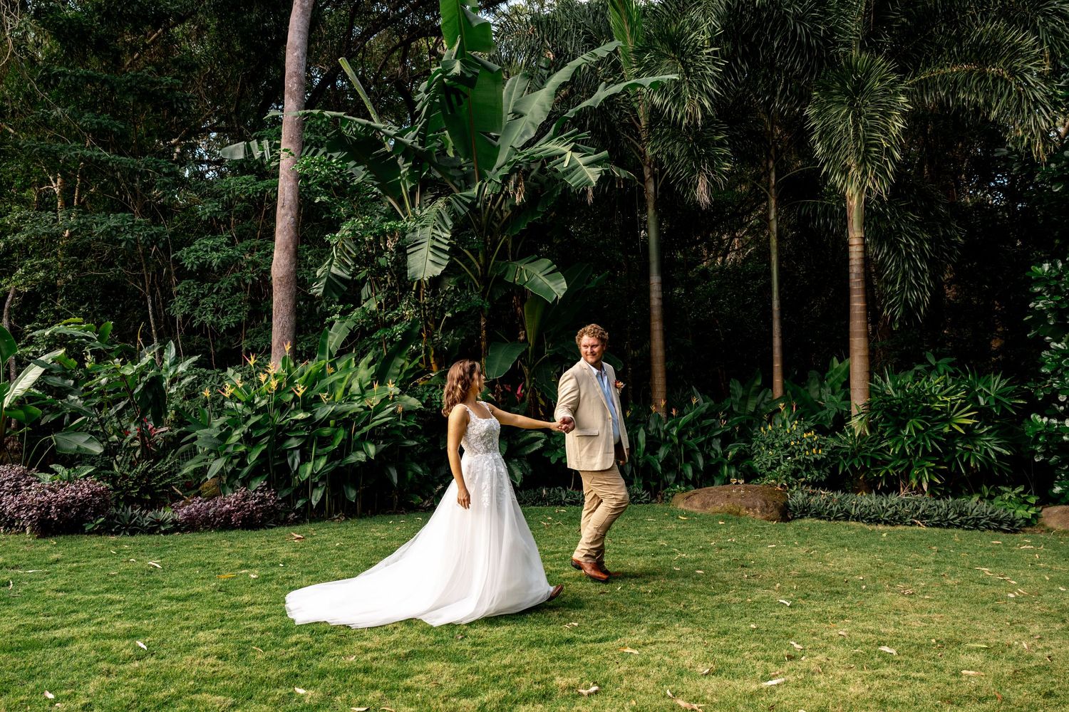 Bride and groom walking hand in hand across green lawn with tropical palms and garden backdrop.
