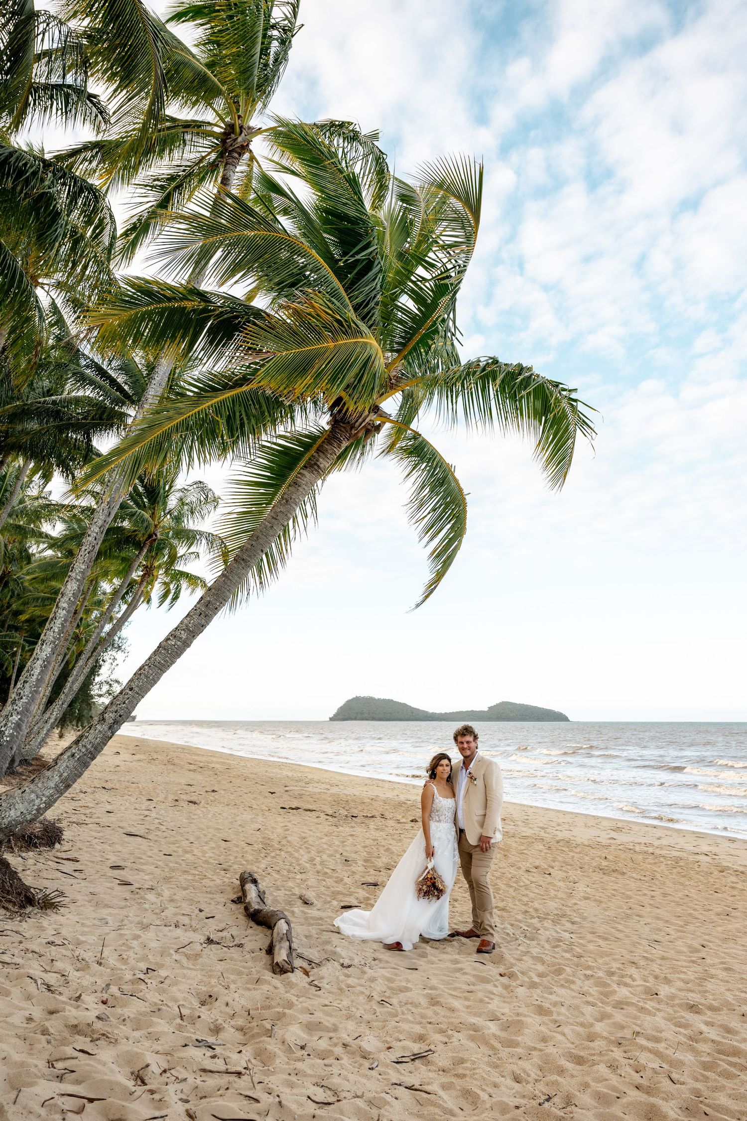 Newlywed couple stands together under a palm tree on a sunny tropical beach with island views.