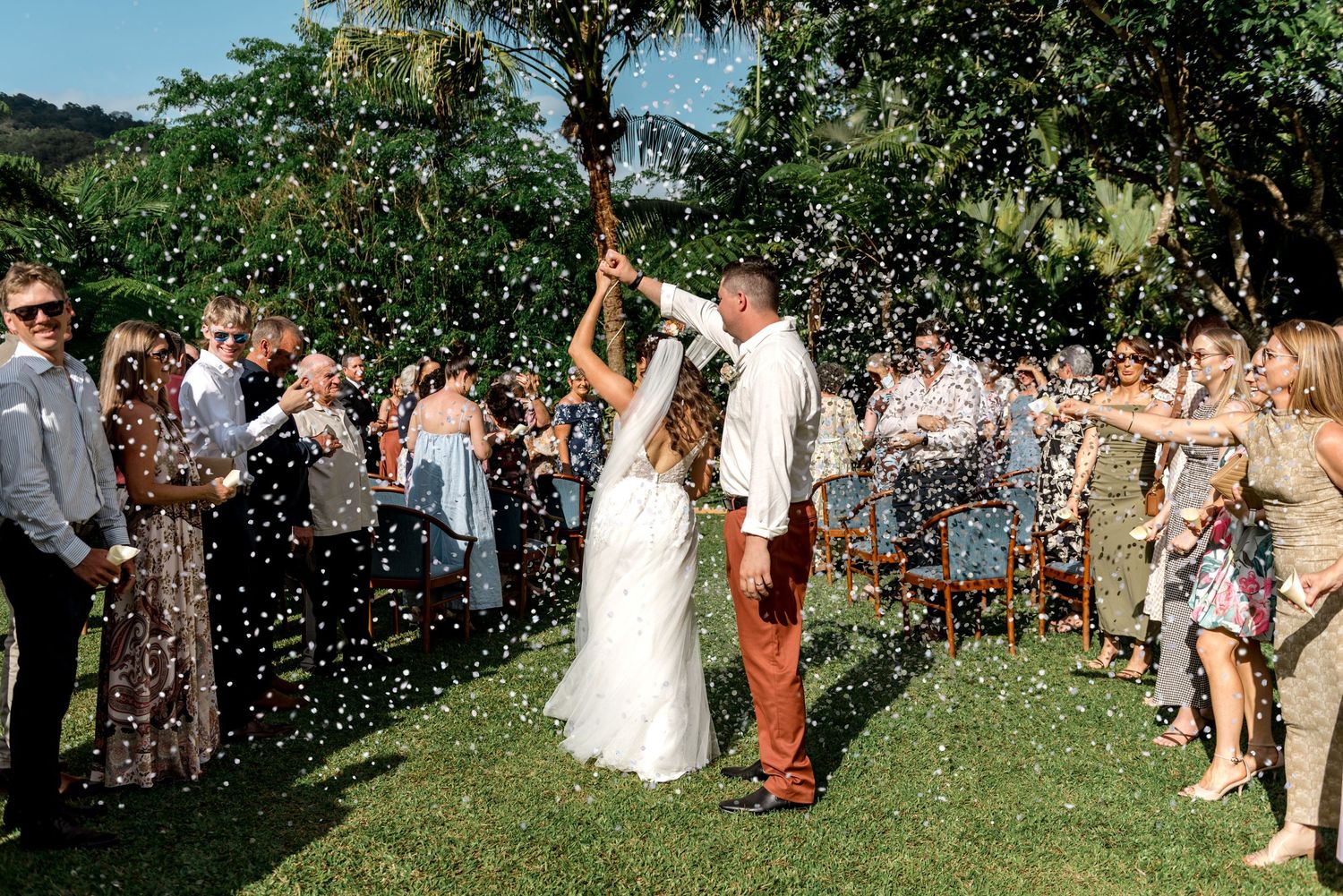Bride and groom walk through confetti shower surrounded by cheering wedding guests outdoors.