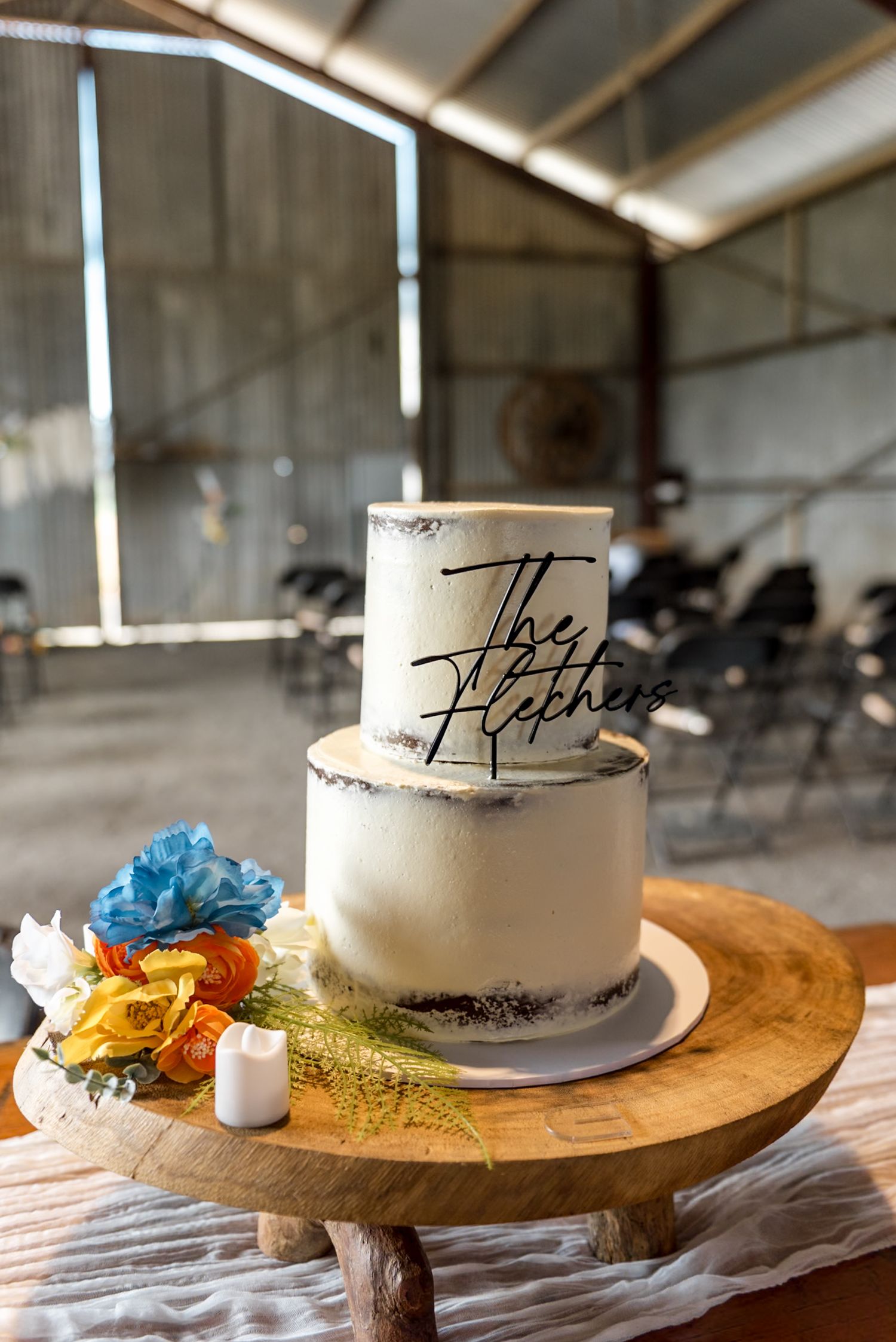 Two-tier semi-naked wedding cake with The Fletchers topper on rustic wooden stand with colorful flowers.