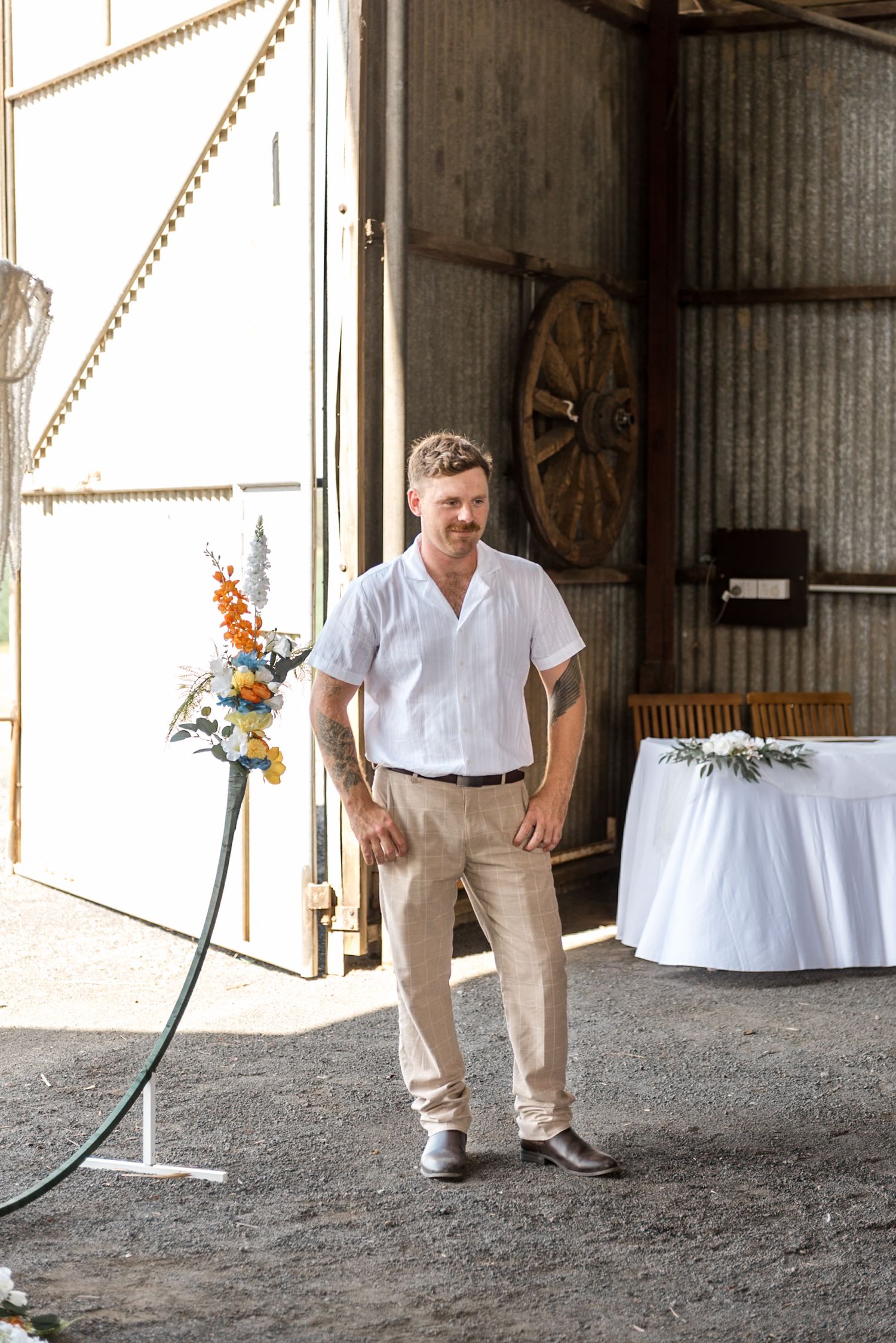 A smiling groom in a white linen shirt and khaki pants stands in a rustic barn wedding venue with floral decorations.