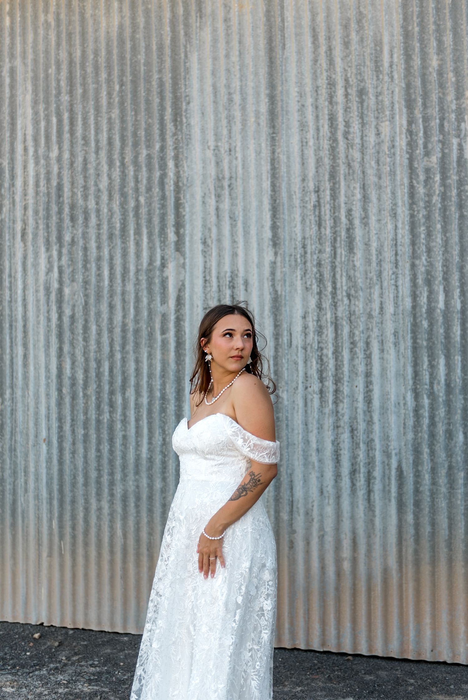 Elegant bride in off-shoulder lace wedding gown poses gracefully against a rustic corrugated metal wall.