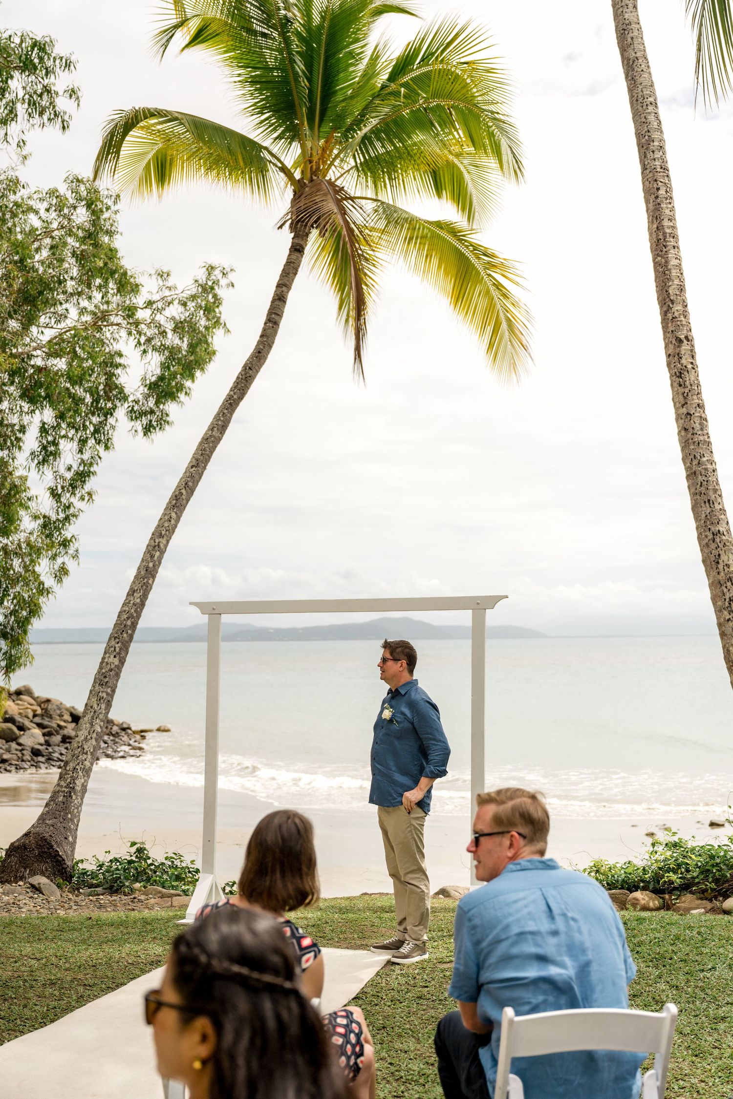 Groom stands at white wedding arch framed by palm trees and ocean backdrop at Little Cove, Port Douglas.