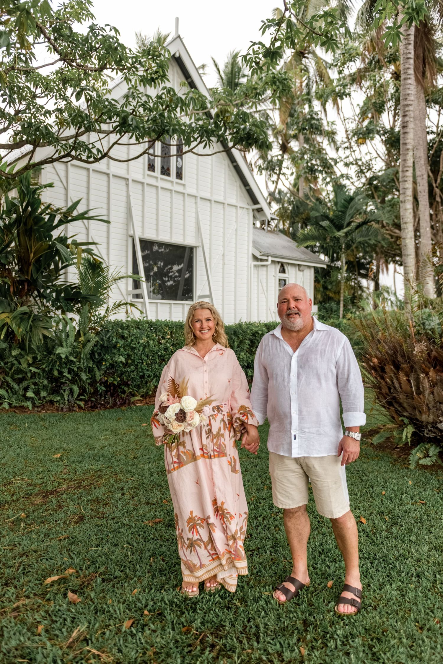 Smiling couple holding hands and a bouquet in front of a white heritage chapel.