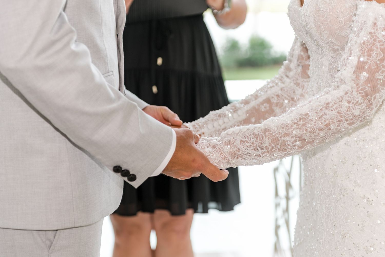 Bride and groom holding hands during wedding ceremony, close-up of lace bridal gown and gray suit.