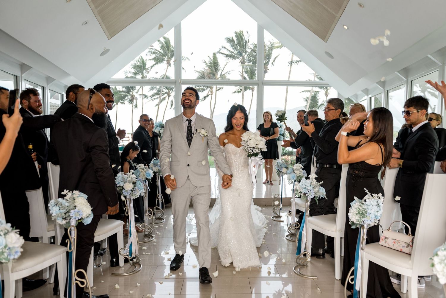 Newlyweds walk down aisle in modern beachfront chapel surrounded by guests dressed in black attire.