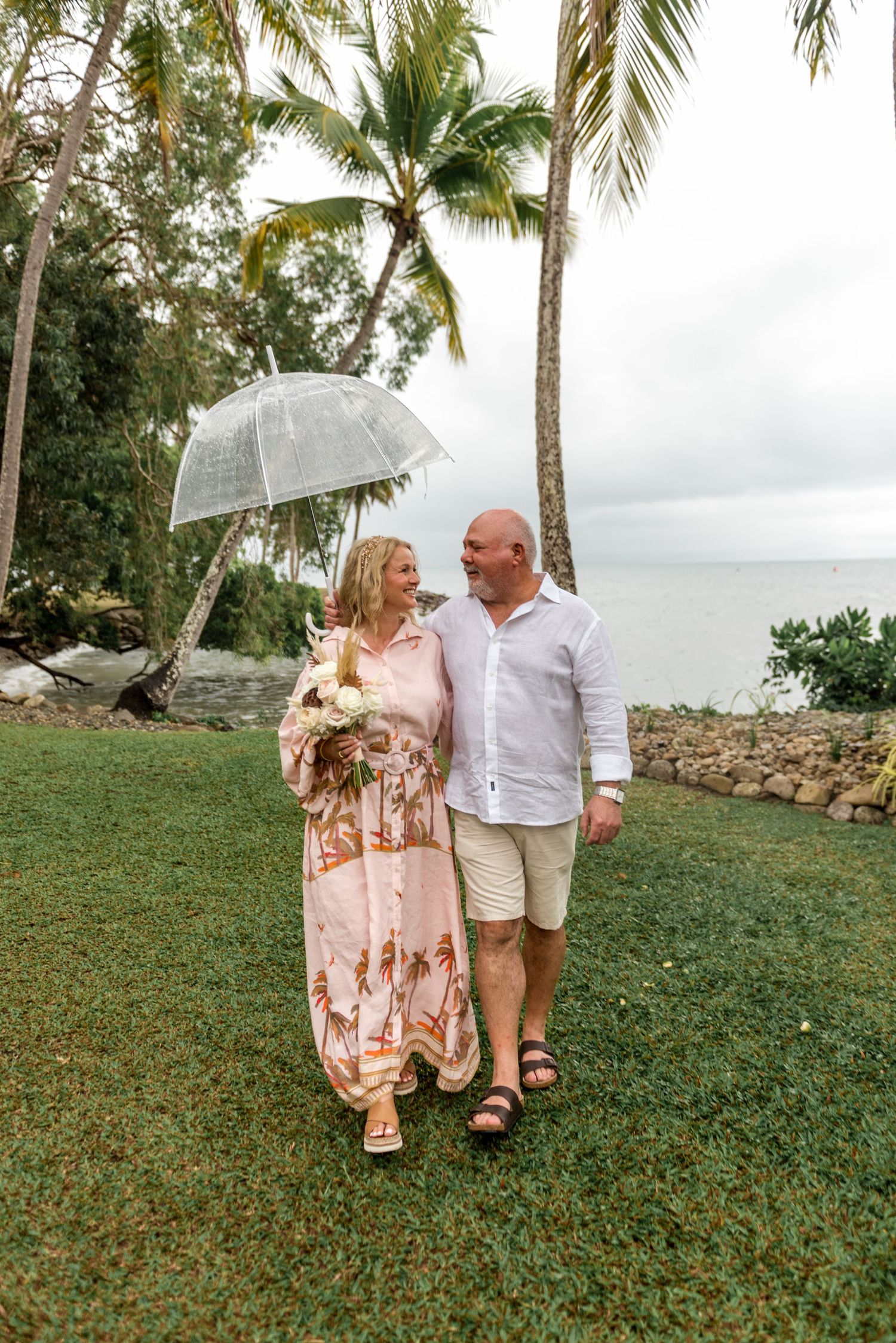 Smiling couple strolling through lush green lawn under a clear umbrella surrounded by tropical palm trees.