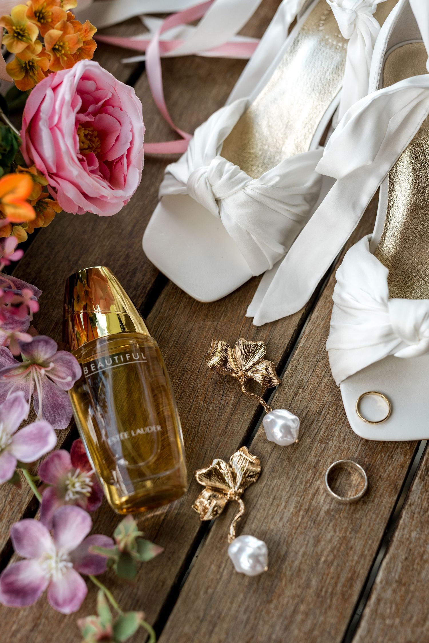 Wedding flat lay featuring Estee Lauder Beautiful perfume, white heels, gold earrings, and rings with flowers.