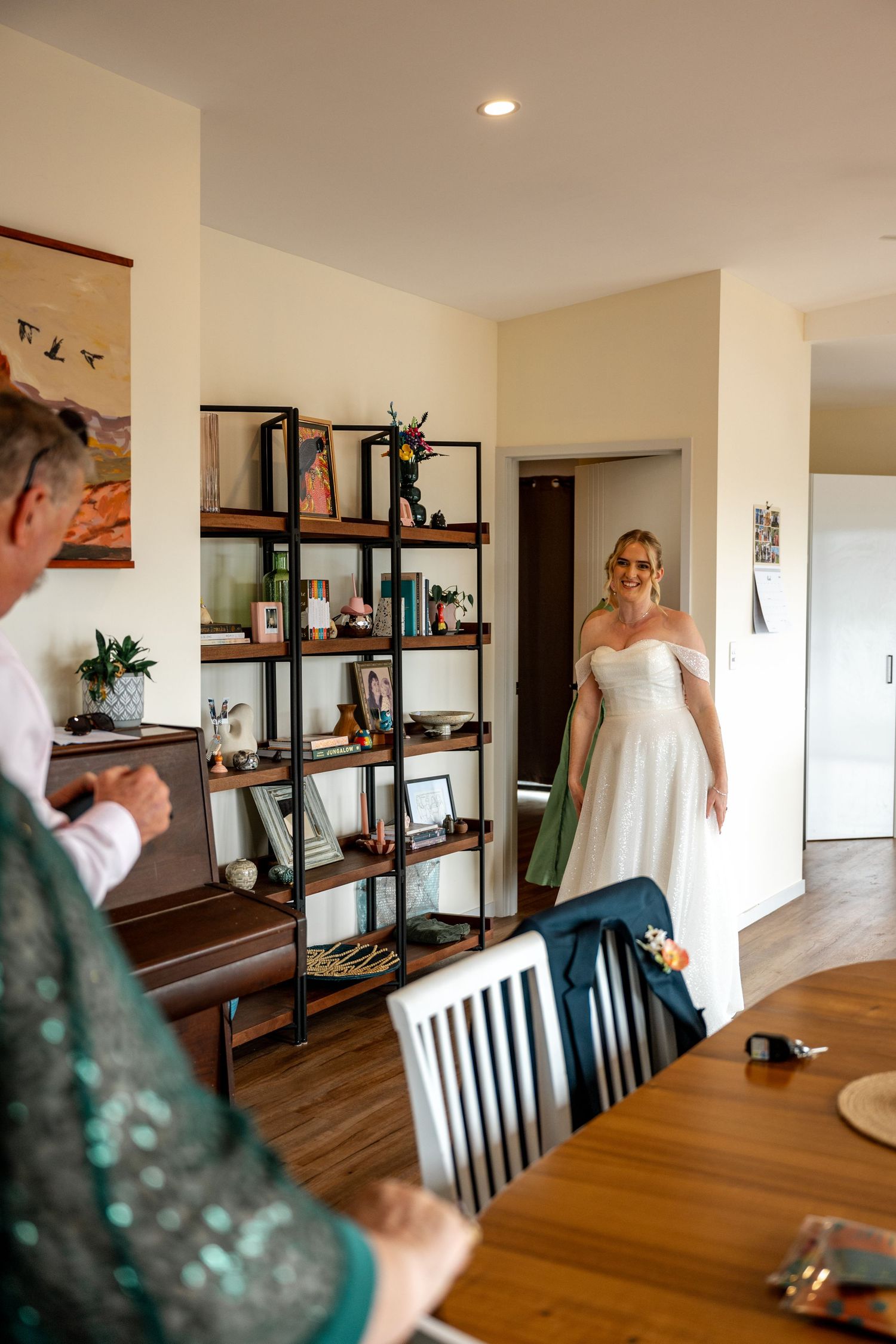Blonde bride in off-shoulder white gown smiling as she enters room during wedding morning prep.