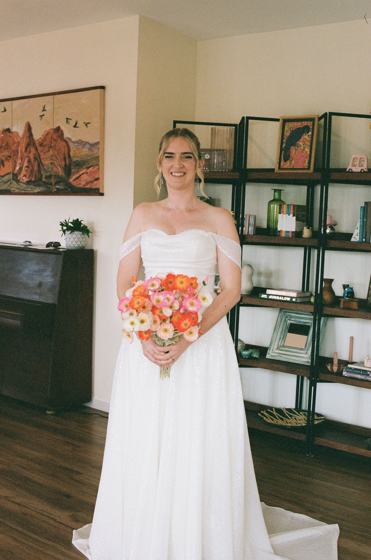 Radiant bride in off-shoulder white gown holds vibrant orange and pink bouquet while smiling indoors before ceremony.