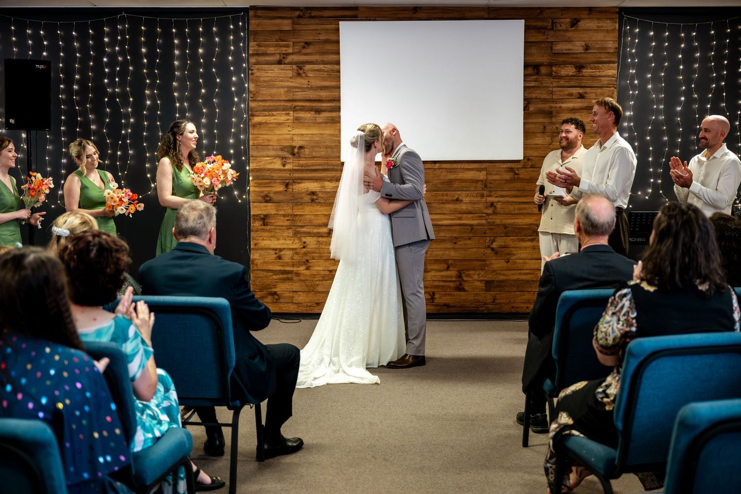 Bride and groom share first kiss at altar while bridesmaids in green dresses look on.