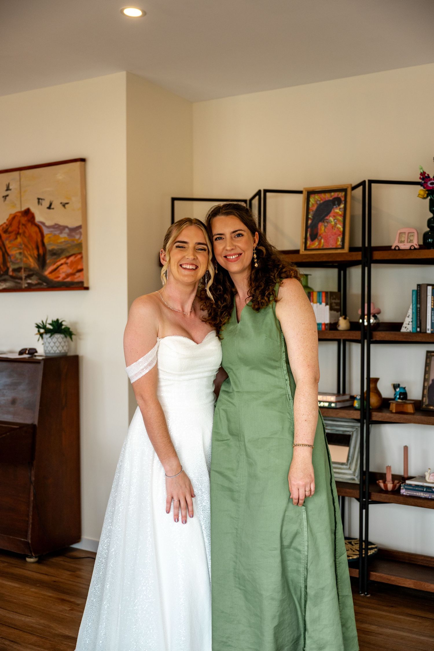 Smiling bride in white off-shoulder gown poses with bridesmaid in green dress before wedding ceremony.