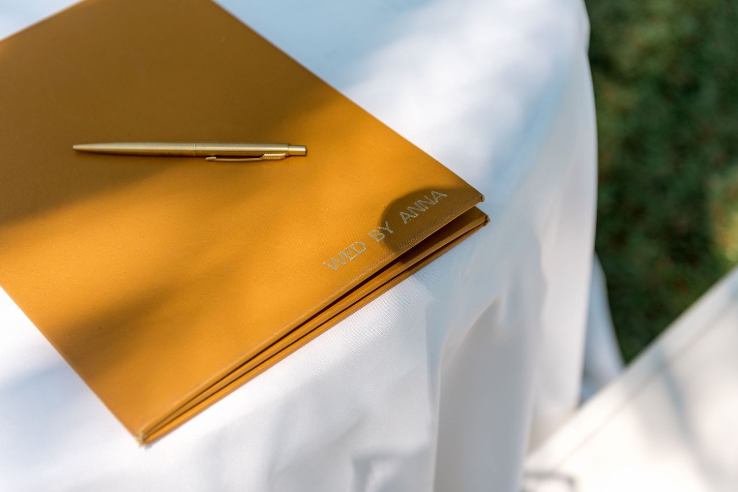 Gold wedding guest book with pen on a white cloth-covered table at an outdoor ceremony.
