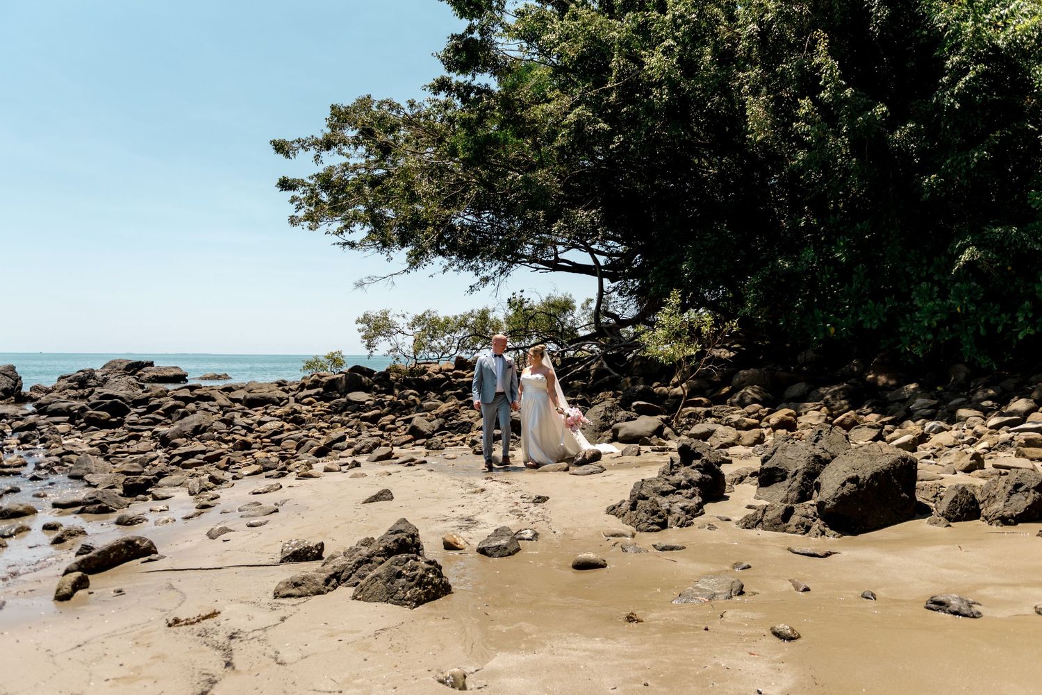 Bride and groom standing together on a sandy beach surrounded by rocks and tropical trees.