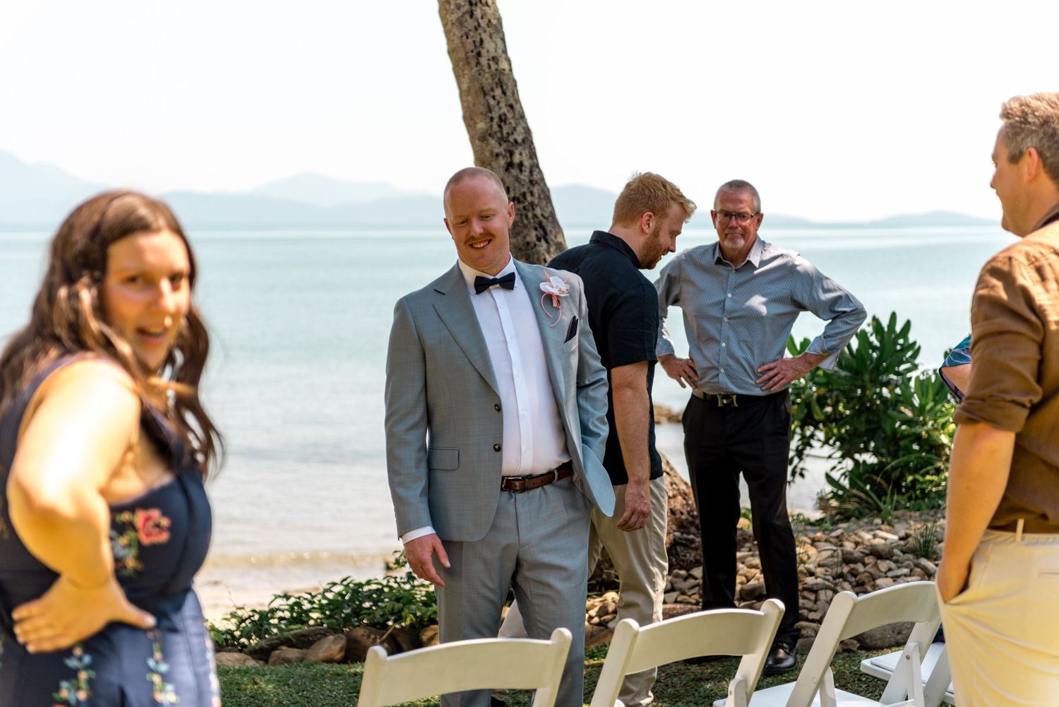 Groom in gray suit and bow tie smiles at guests near palm tree at beachside wedding.