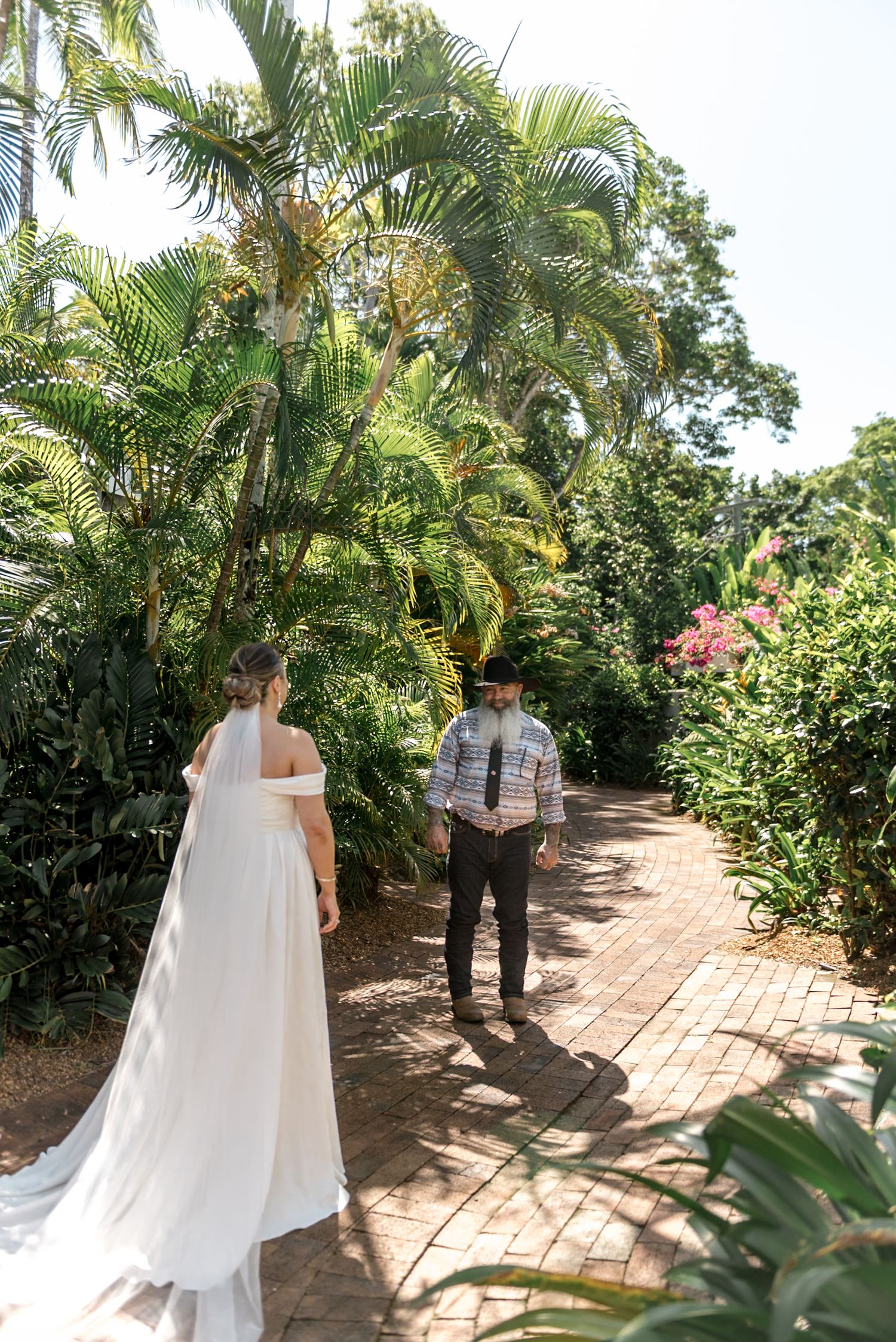 Bride in white gown walks toward groom on brick path surrounded by lush tropical garden greenery.