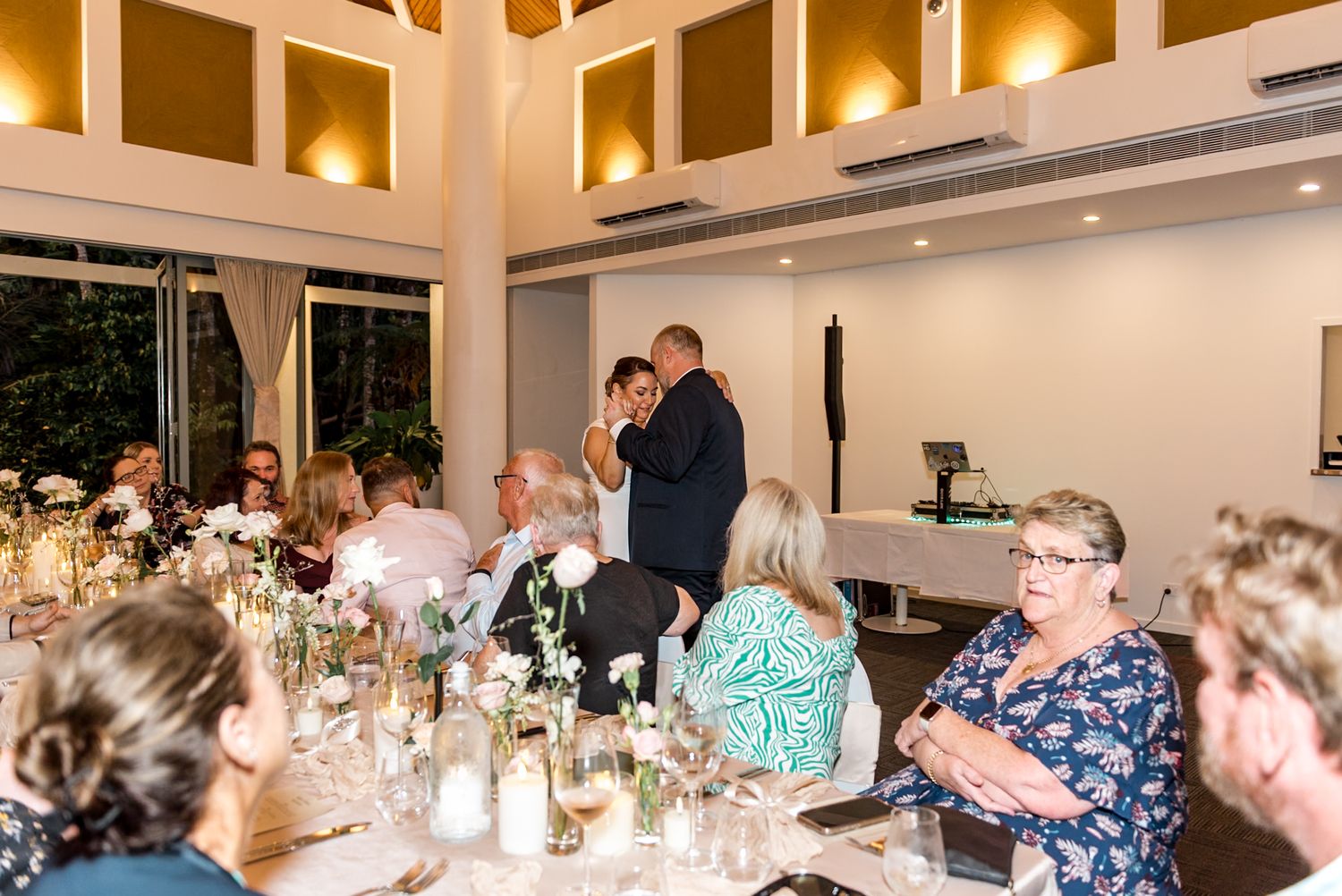 Wedding couple embrace on dance floor while guests enjoy candlelit reception dinner tables.