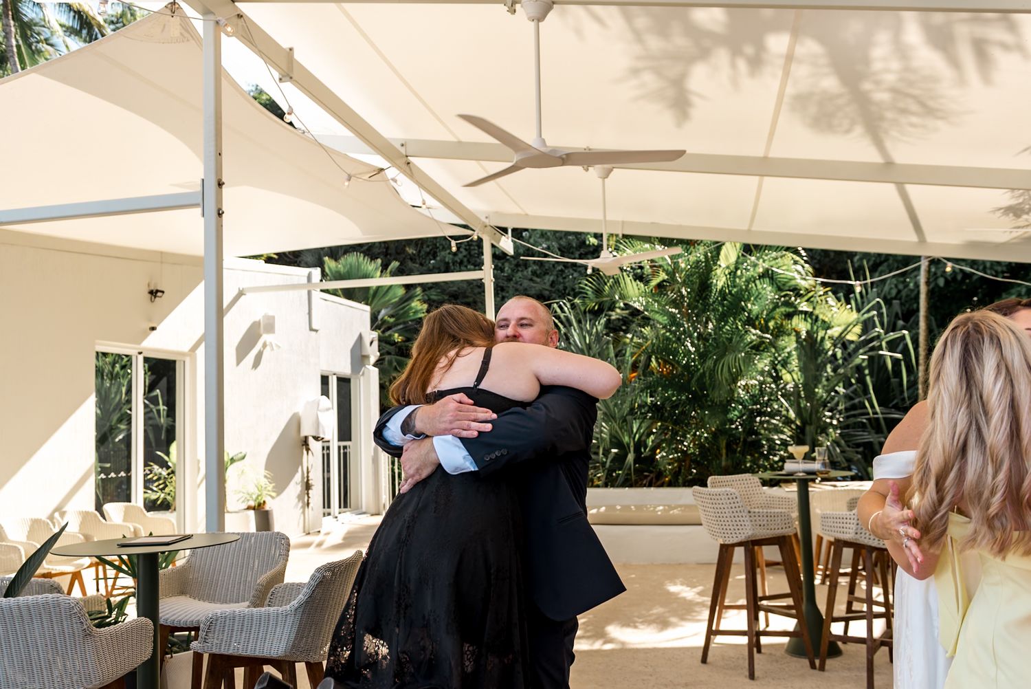 Two guests share a warm hug at an outdoor wedding reception under a white canopy.