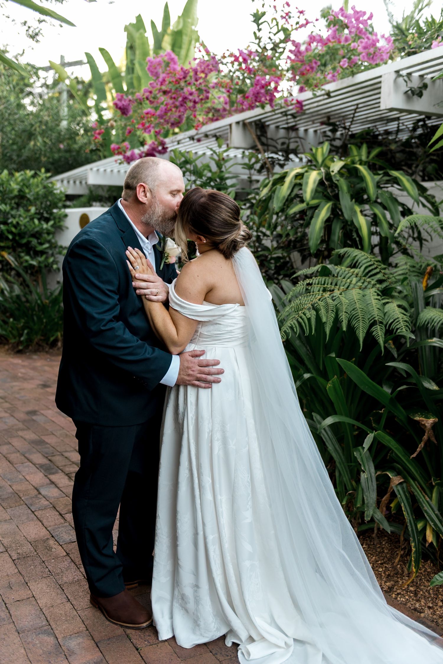 Groom in navy suit kissing bride in off-shoulder white gown in lush tropical garden.