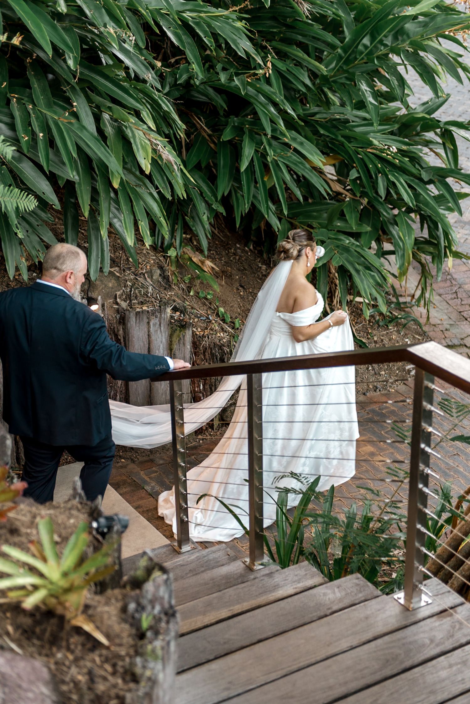 Bride in flowing white gown and veil walks up wooden stairs with groom through lush greenery.