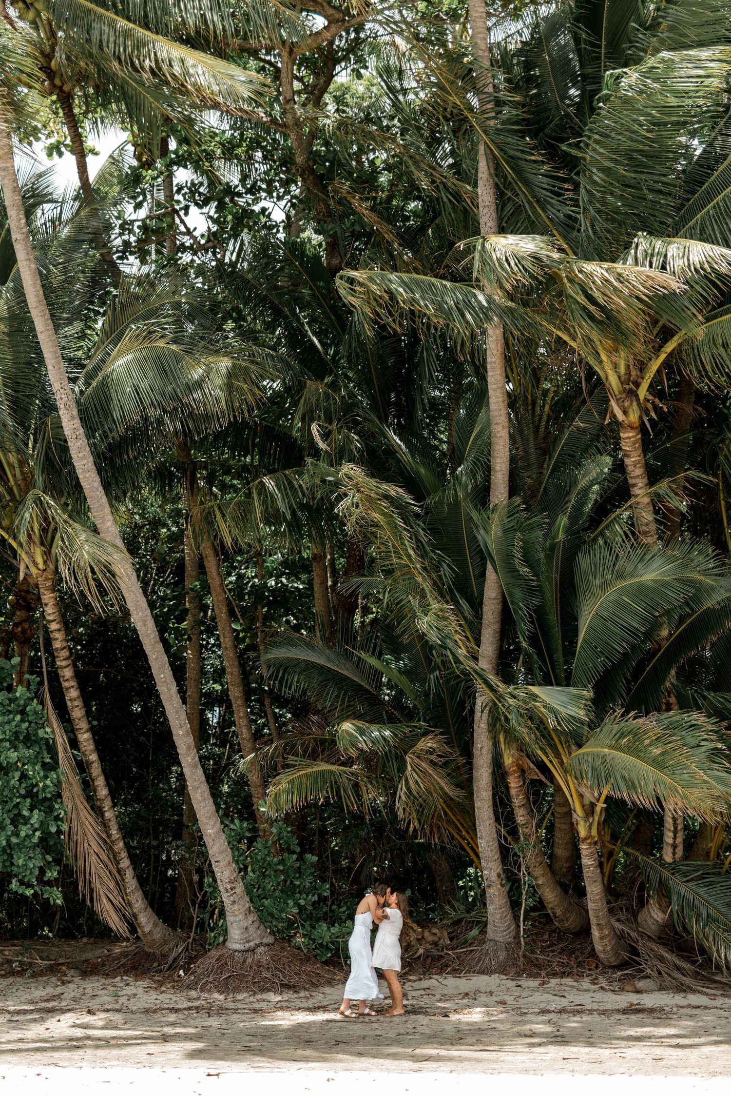 Same-sex couple sharing a romantic kiss on a sandy beach surrounded by tall tropical palm trees.