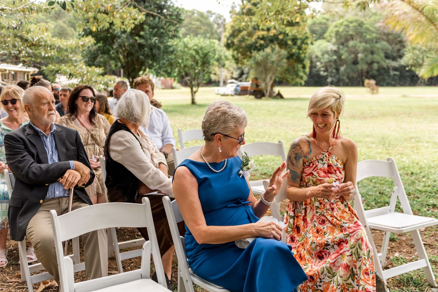 Wedding guests seated on white chairs outdoors, women in colorful dresses chatting before ceremony.