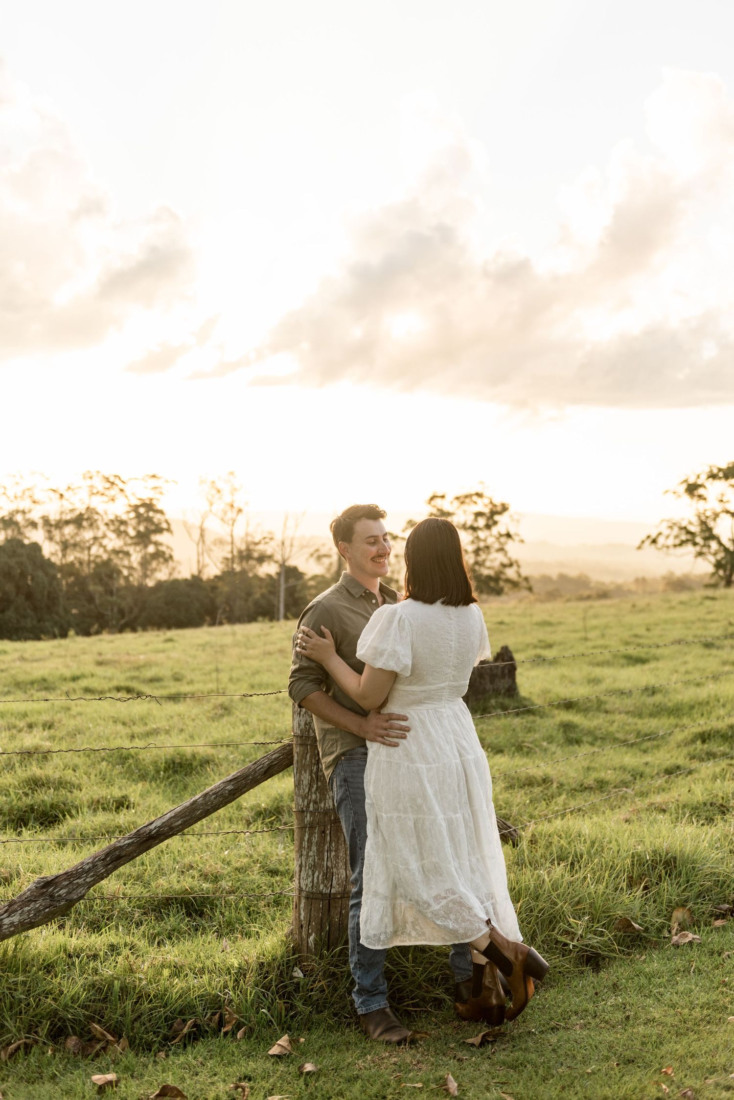 Couple embracing by rustic wooden fence post in golden sunset meadow, woman in white dress.