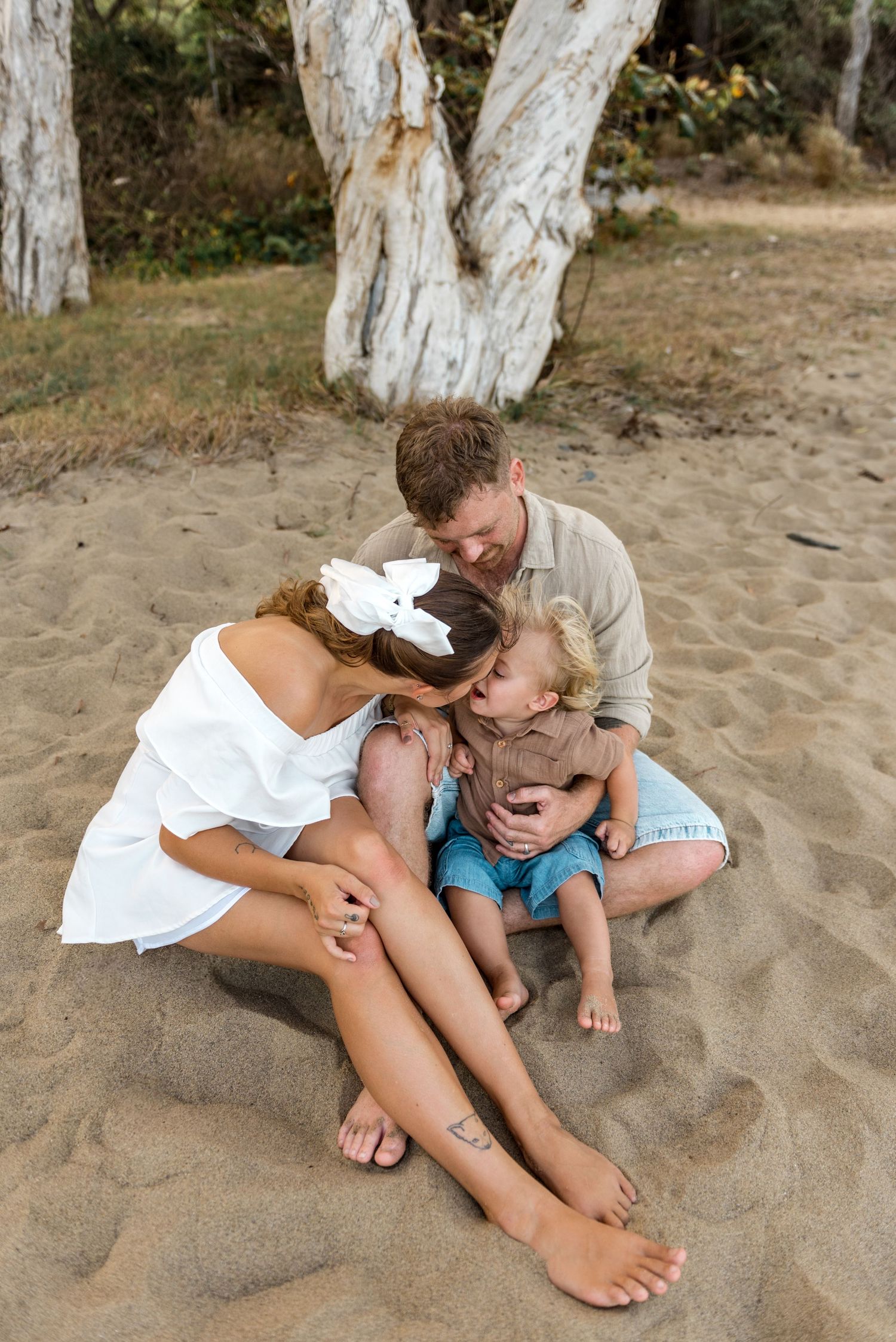 A family cuddles together on the sand as parents kiss their laughing toddler on the beach.