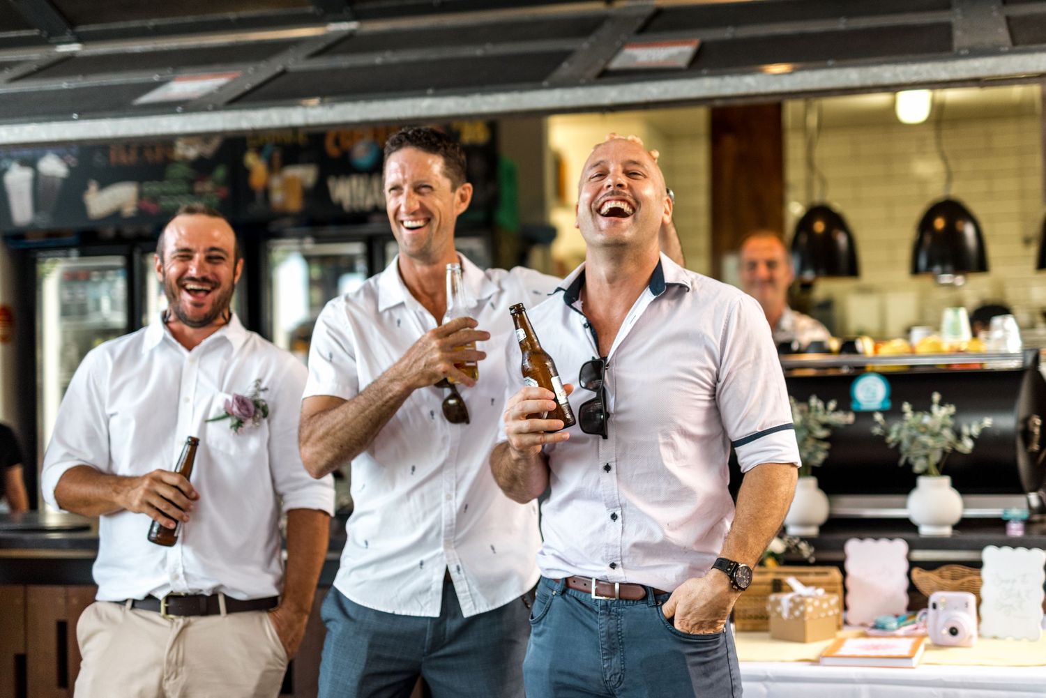 Three men in white shirts laughing and celebrating with beer bottles at wedding reception bar.