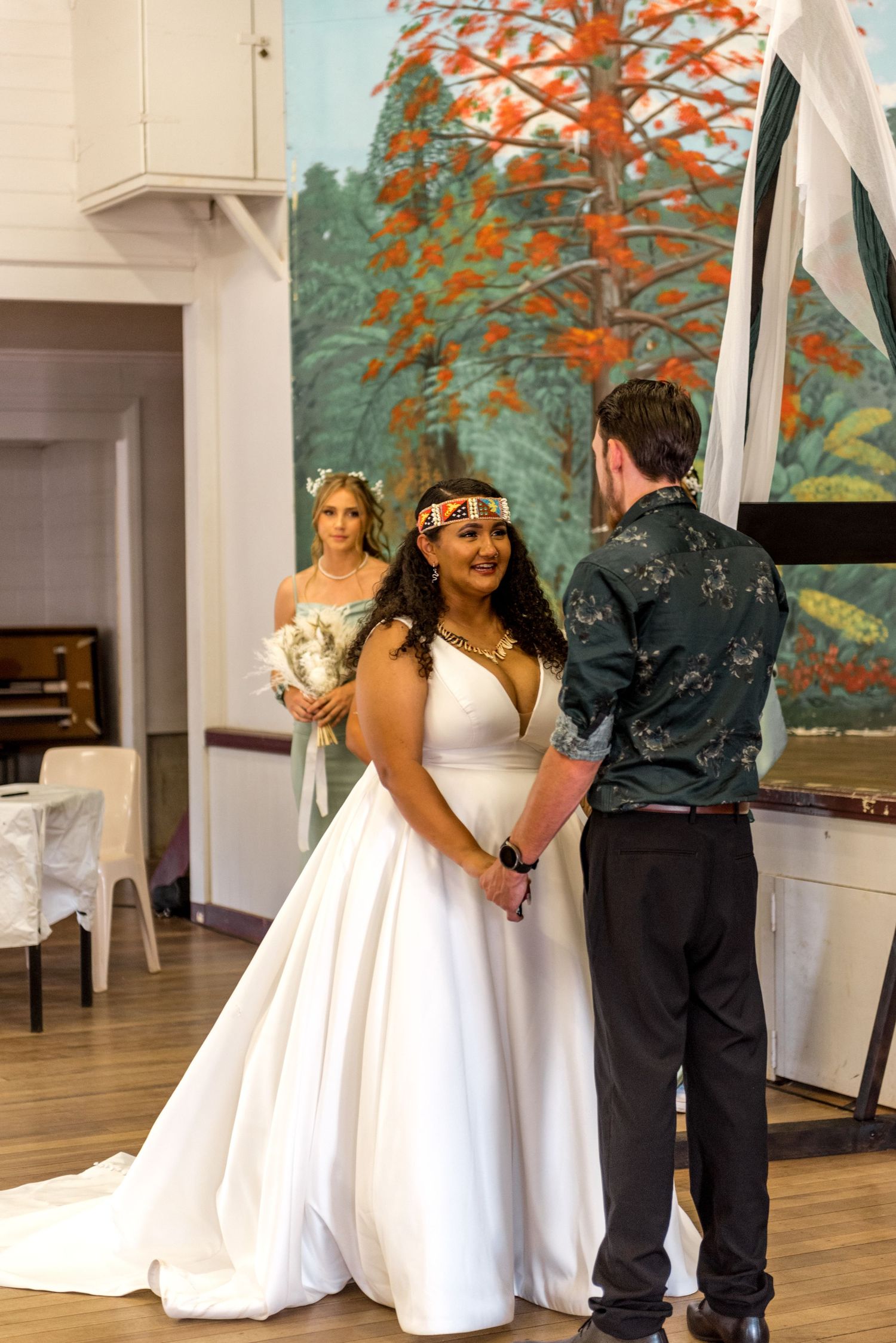 Bride in white gown with beaded headpiece holds hands with groom during indoor wedding ceremony before mural.
