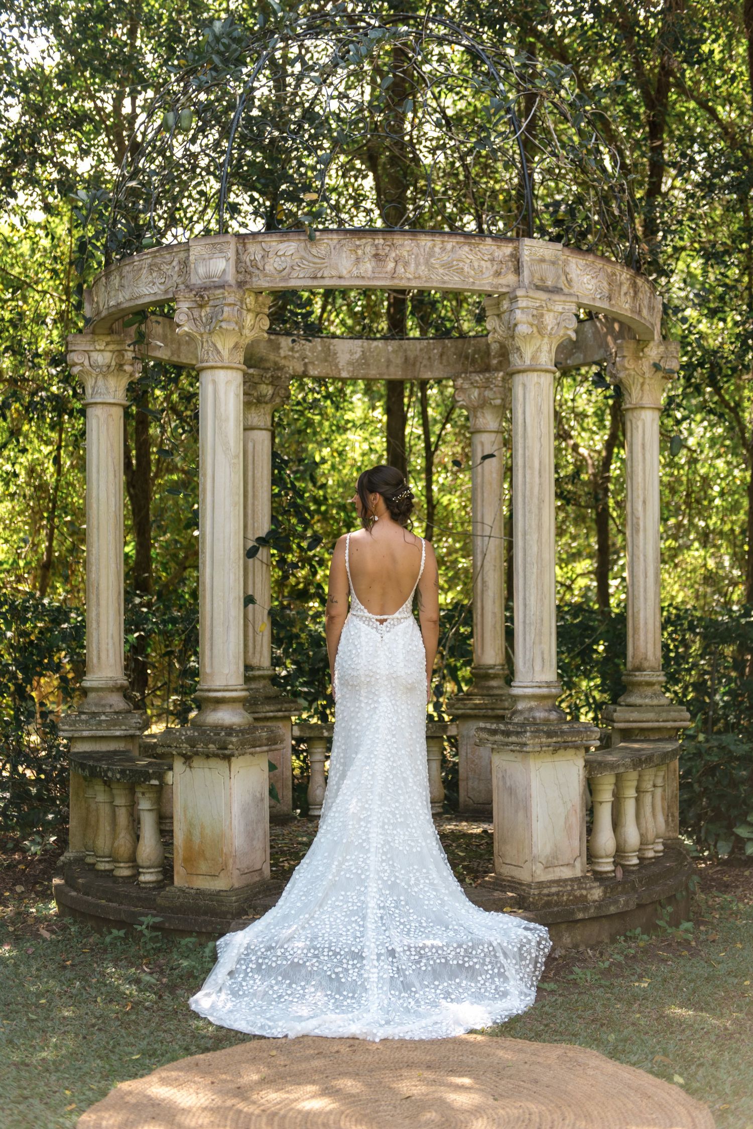 Bride in elegant backless lace wedding gown with long train standing inside ornate stone garden gazebo.